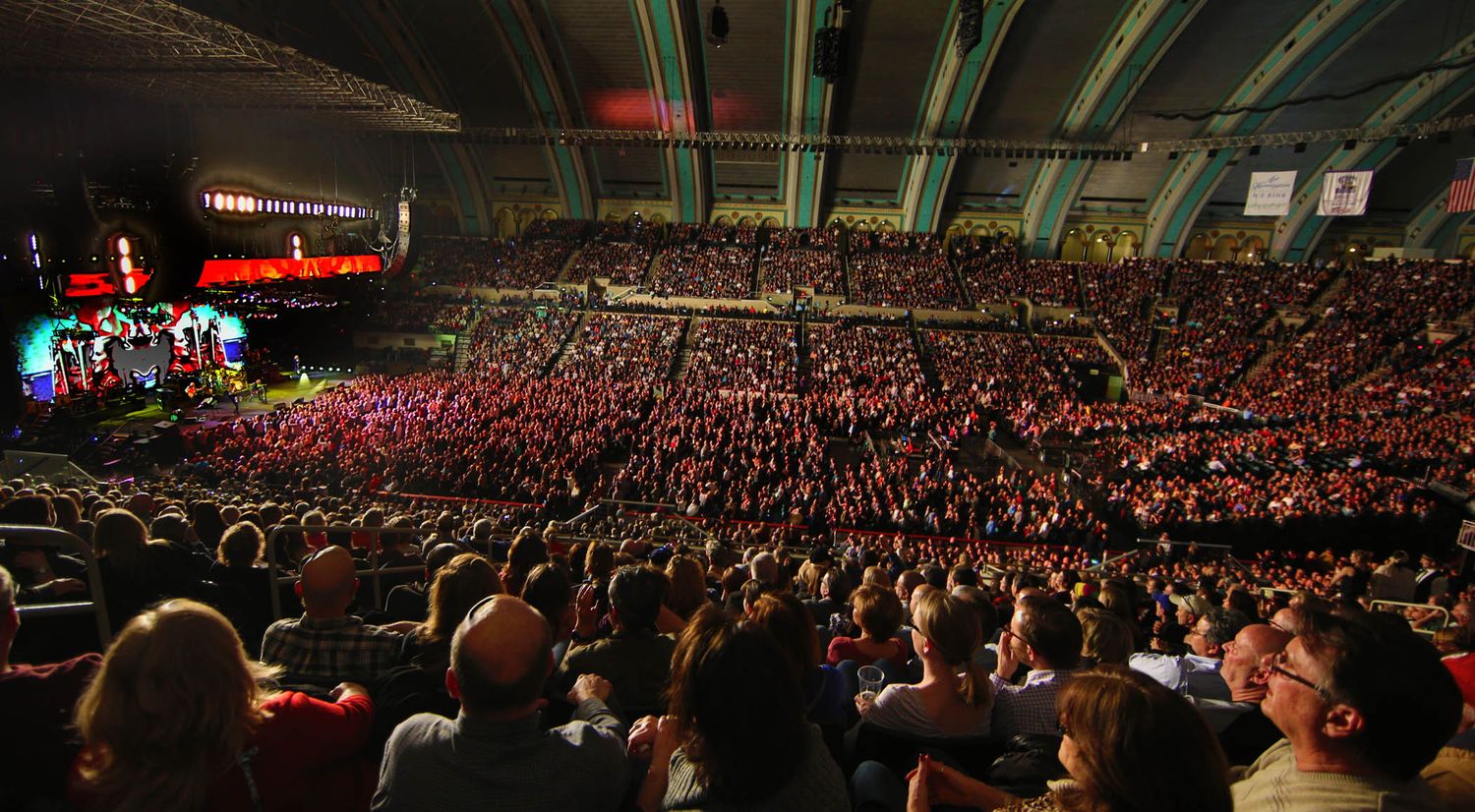 Wide panoramic view of packed concert arena with crowd and stage lighting.