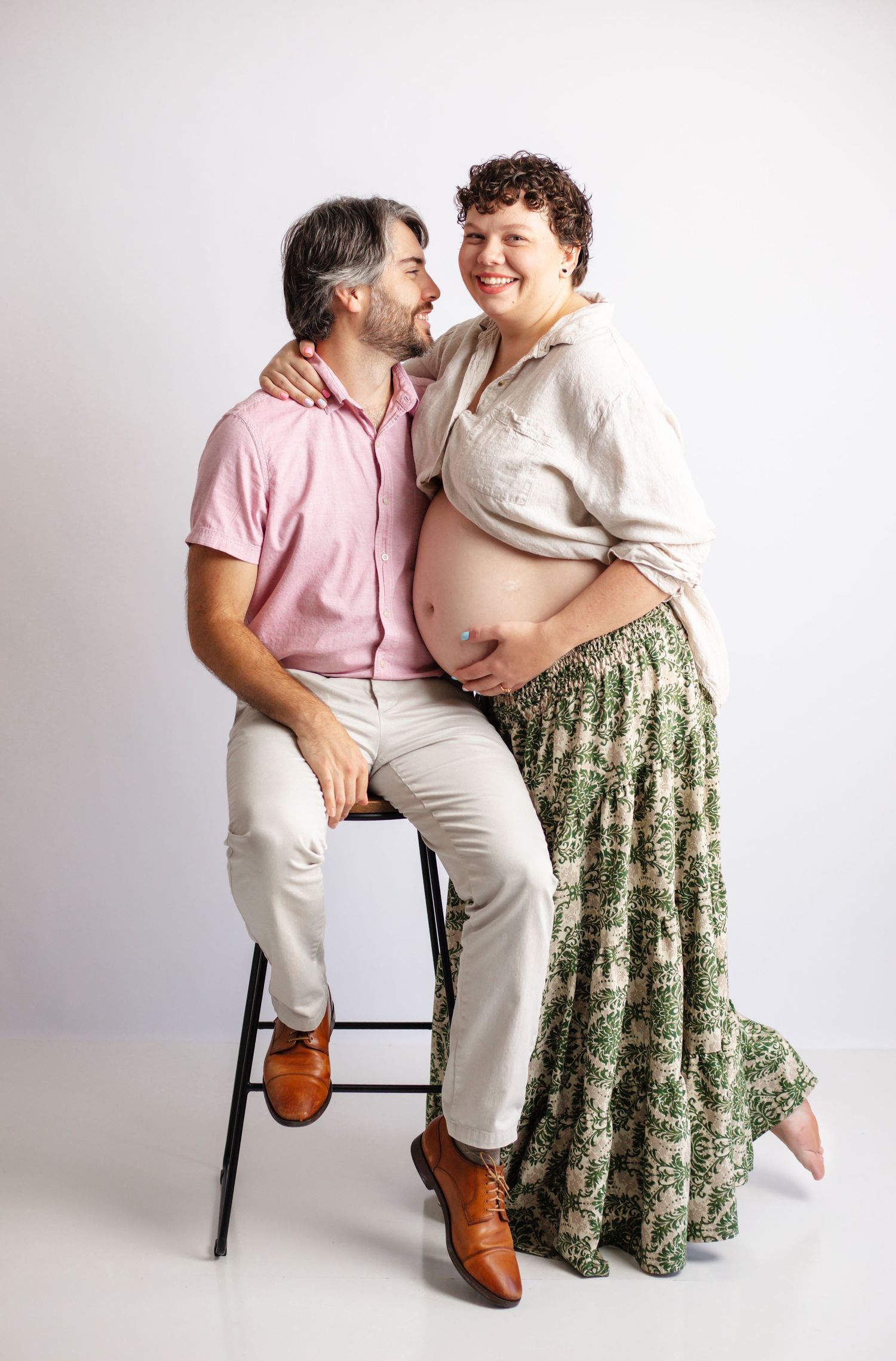 Two people share an intimate moment while sitting together on stools against a white backdrop.
