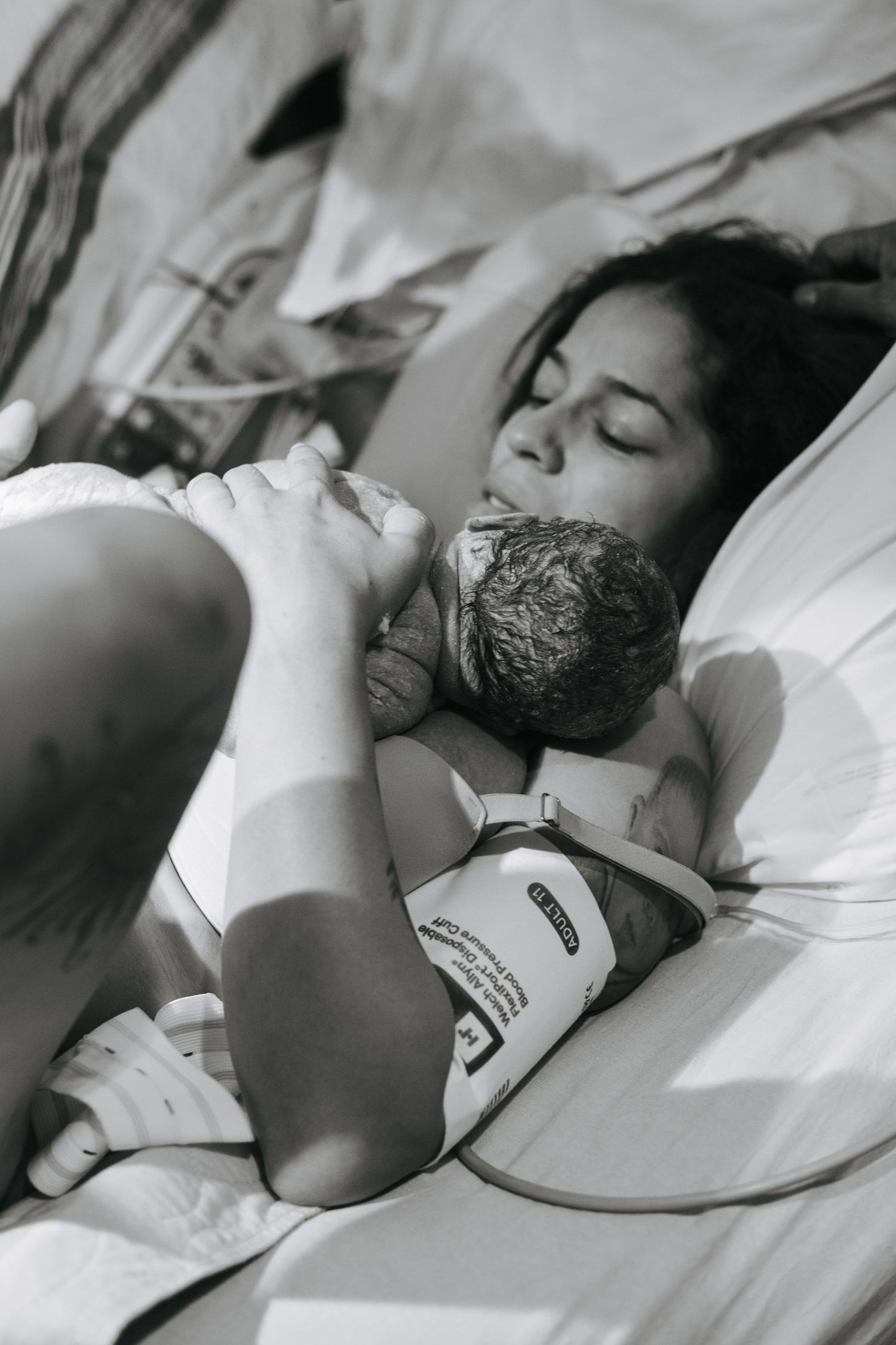 A tender black and white photograph of a mother and newborn baby resting peacefully together on white bedding.