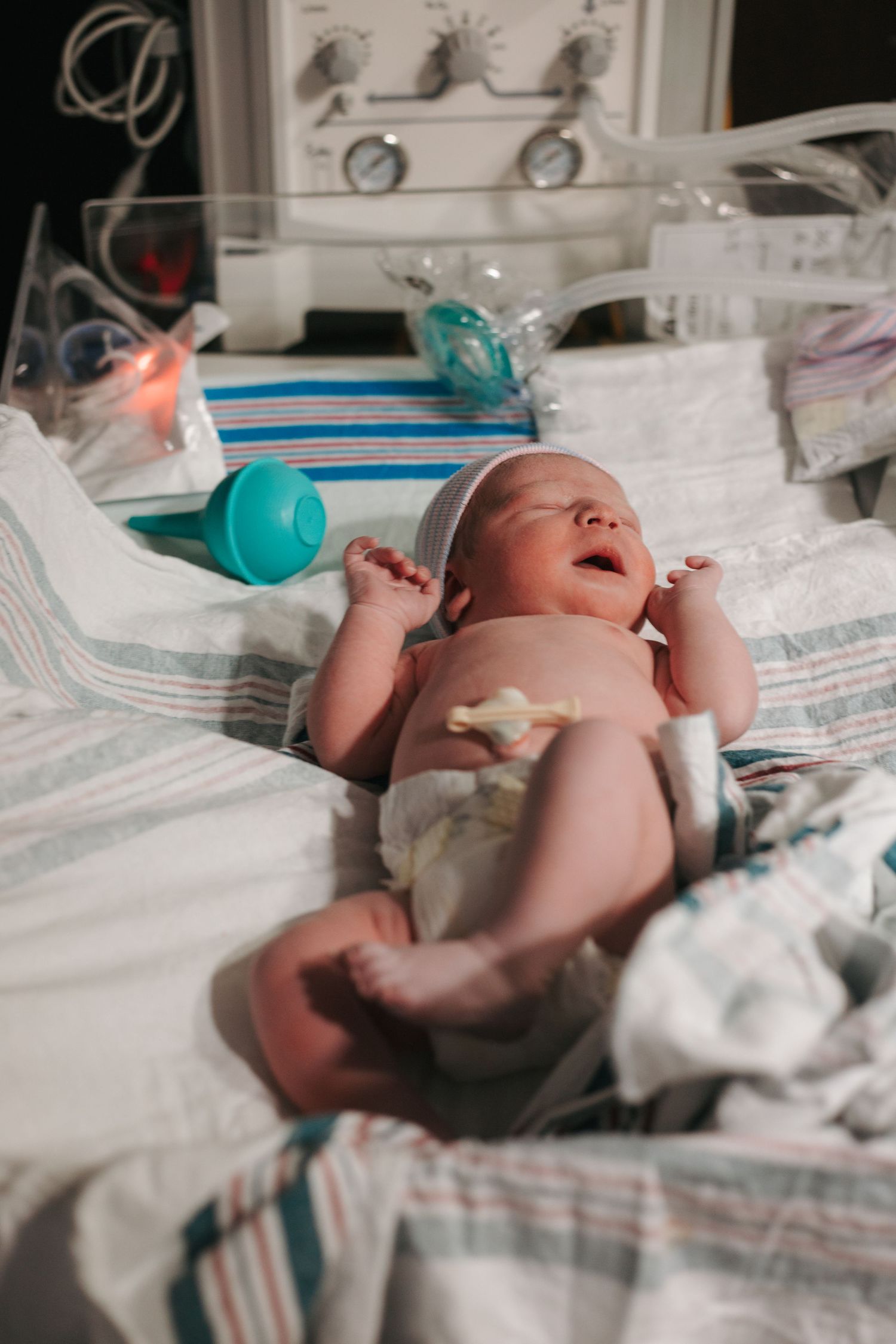 A newborn baby lying on hospital bed with medical equipment in background.