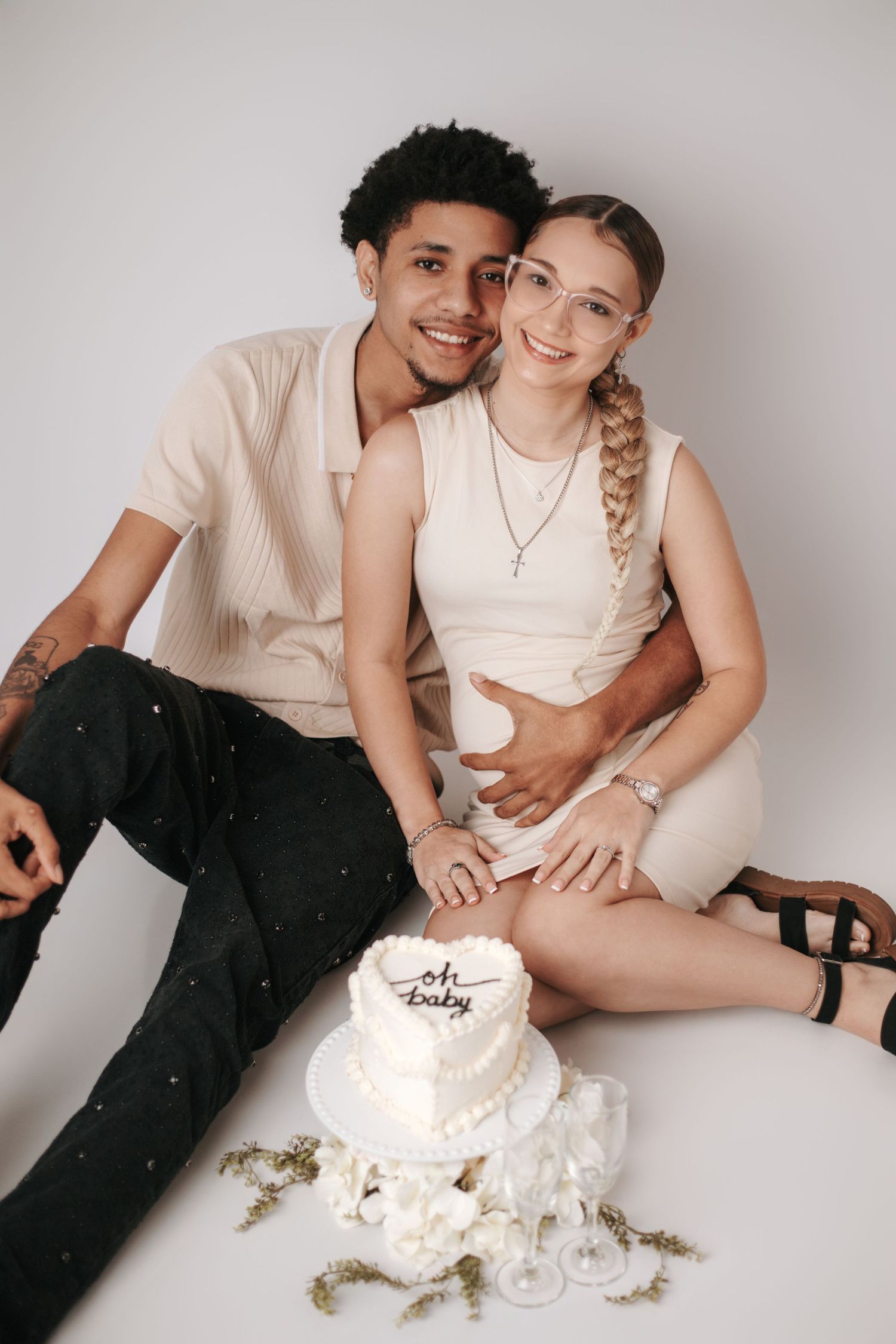 Two people in white outfits sit together playfully on a white background in a romantic photoshoot series.