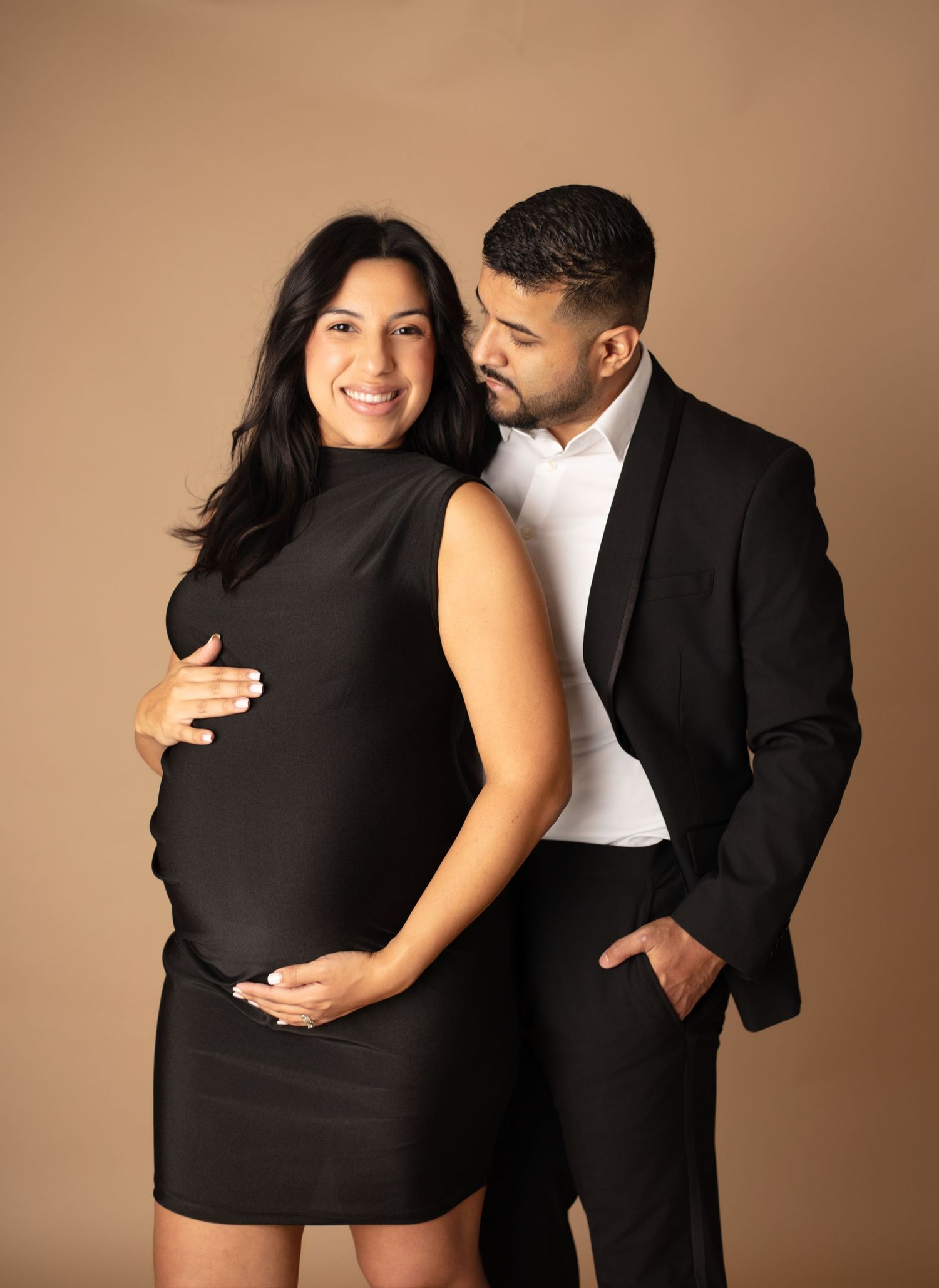 Intimate maternity photo session showing couple in black attire embracing against neutral beige backdrop.