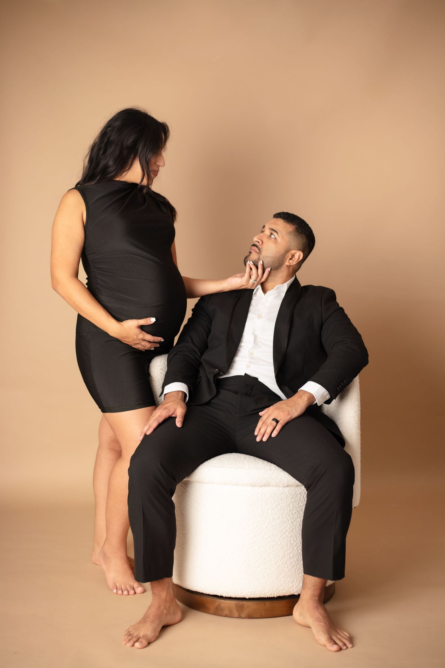 Elegant maternity photo of couple in black attire posing together against beige backdrop with white ottoman.