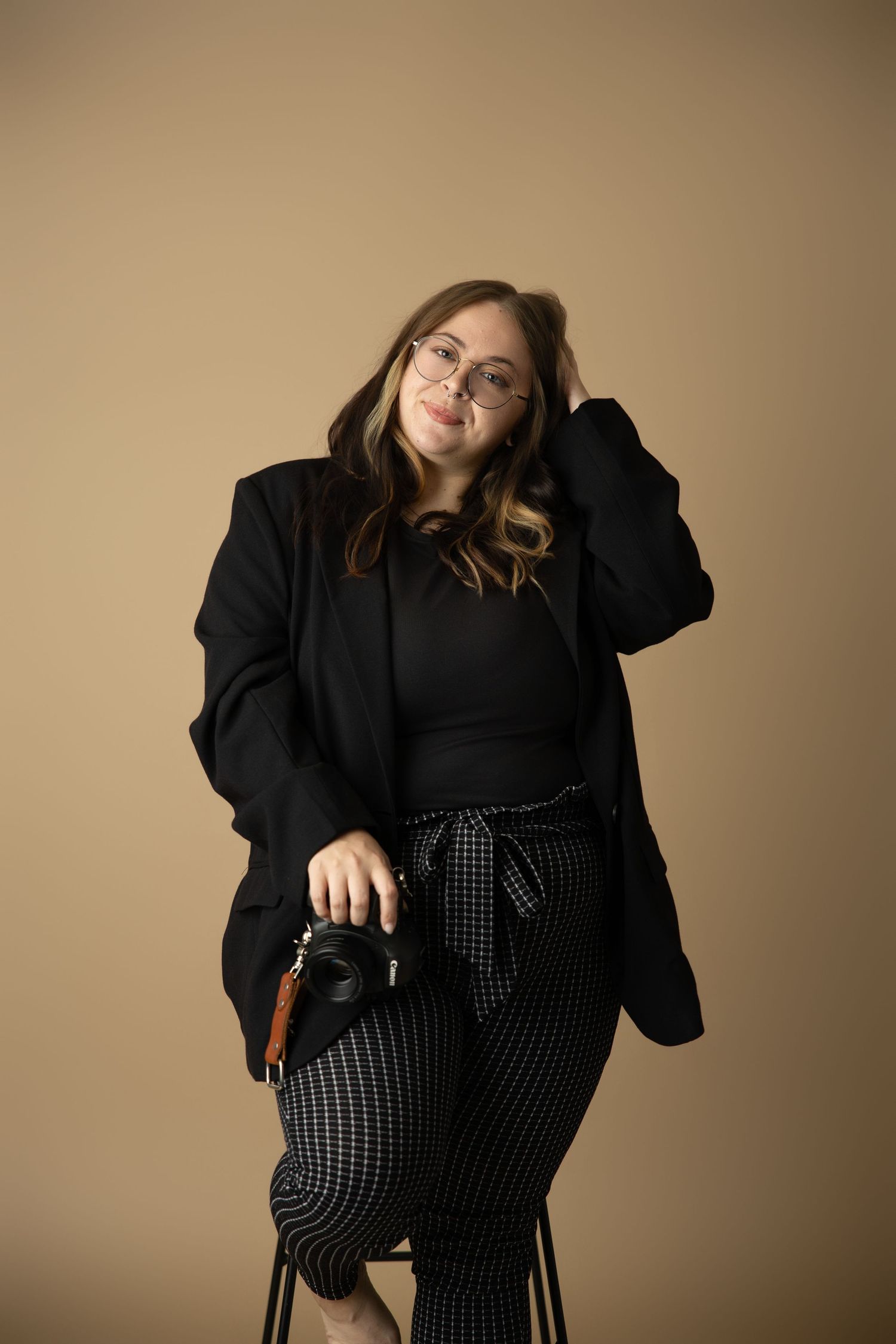 A person in a black blazer and checkered pants poses casually against a neutral backdrop.