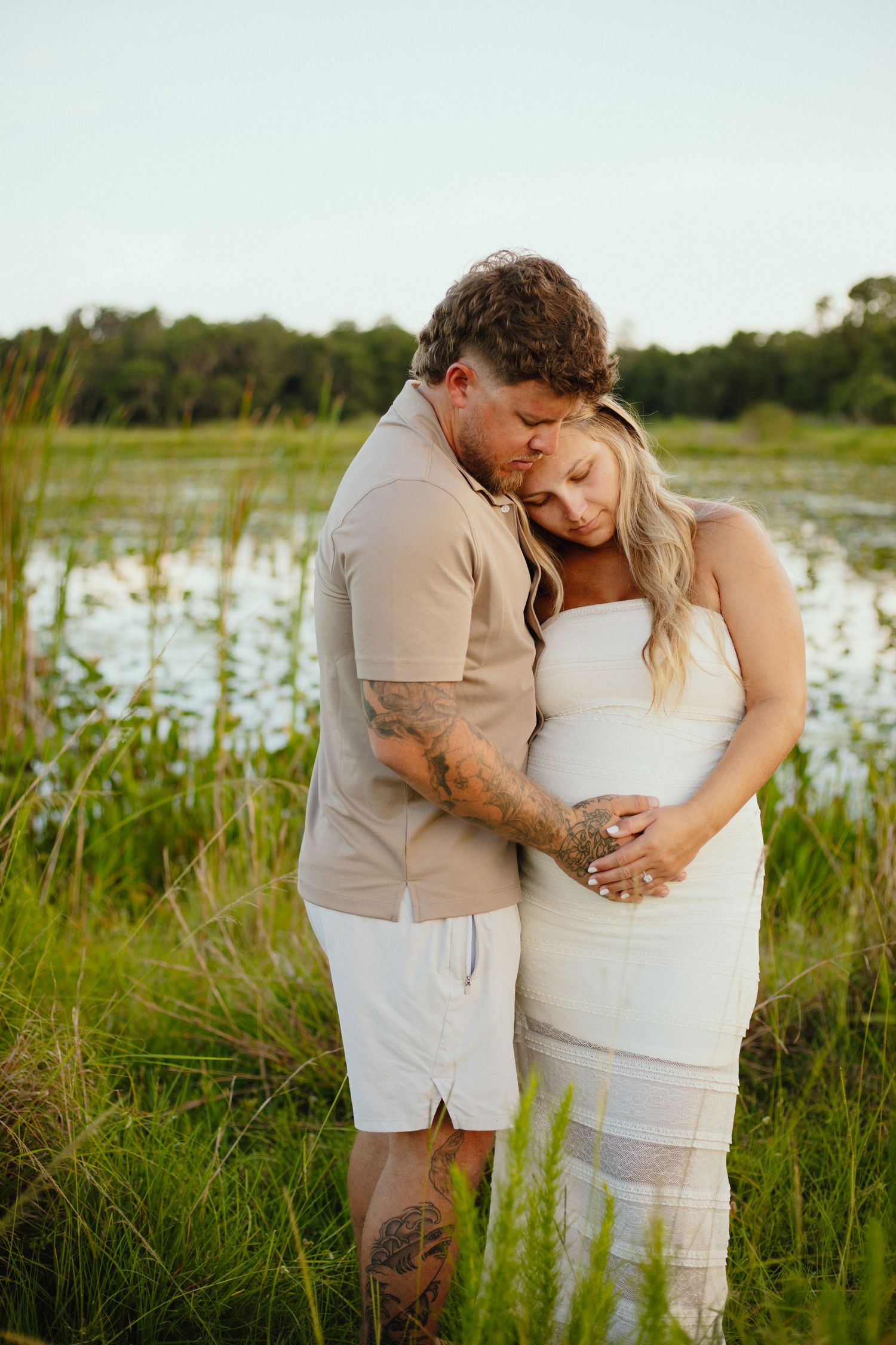 A couple shares an intimate moment by a lake, embracing while standing in tall grass during golden hour.