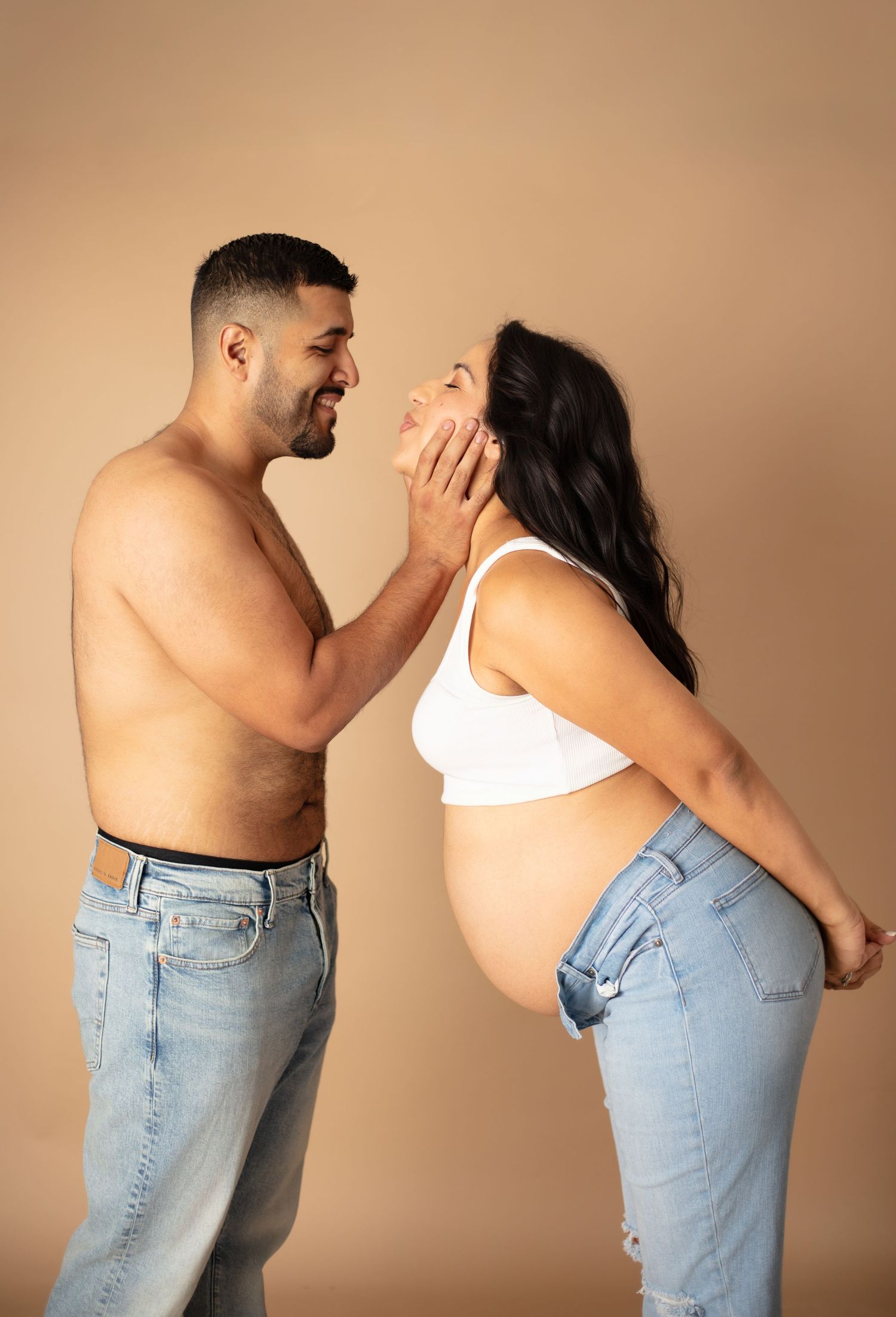 A couple in jeans share a joyful moment during a maternity photoshoot against a neutral brown backdrop.