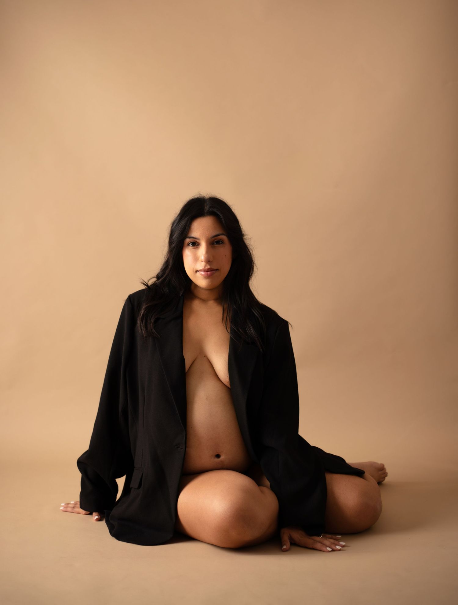 Artistic portrait of a person in an oversized black blazer sitting against a neutral beige background.