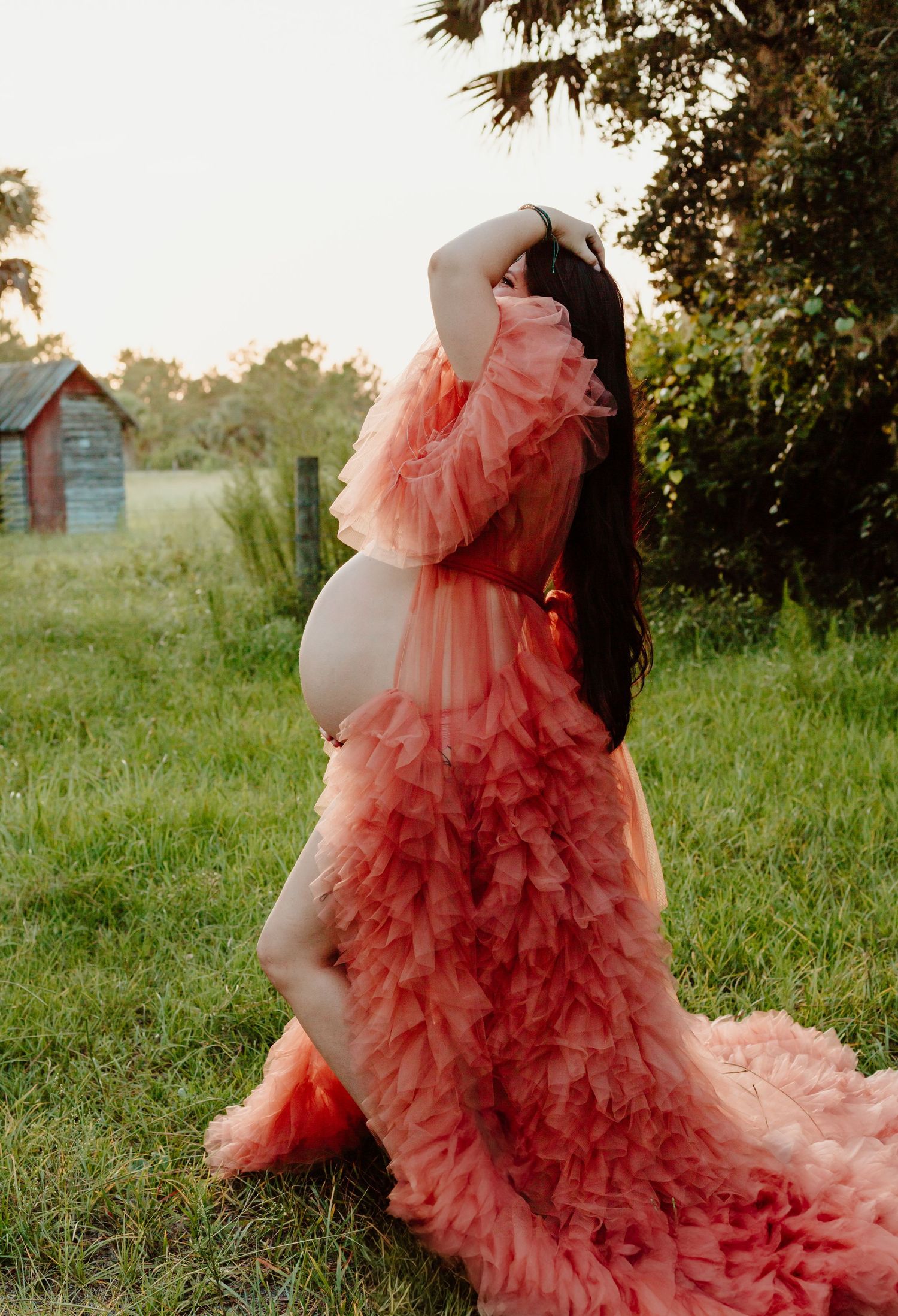 A pregnant woman in a flowing coral ruffled gown poses in a garden at sunset.