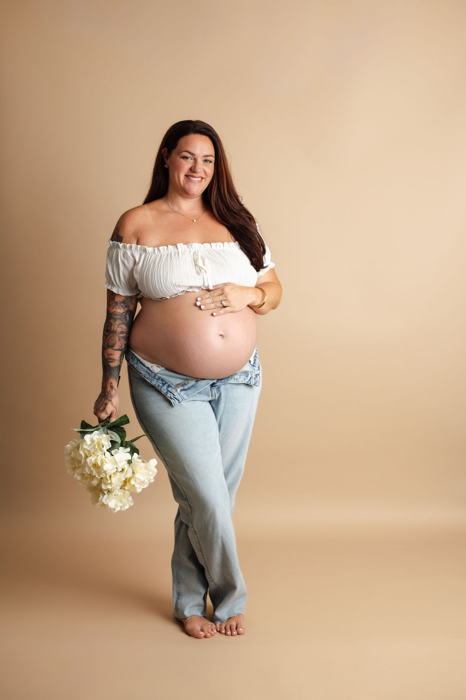 A maternity photo series showing a pregnant woman in light denim and white top holding white flowers against a beige backdrop.