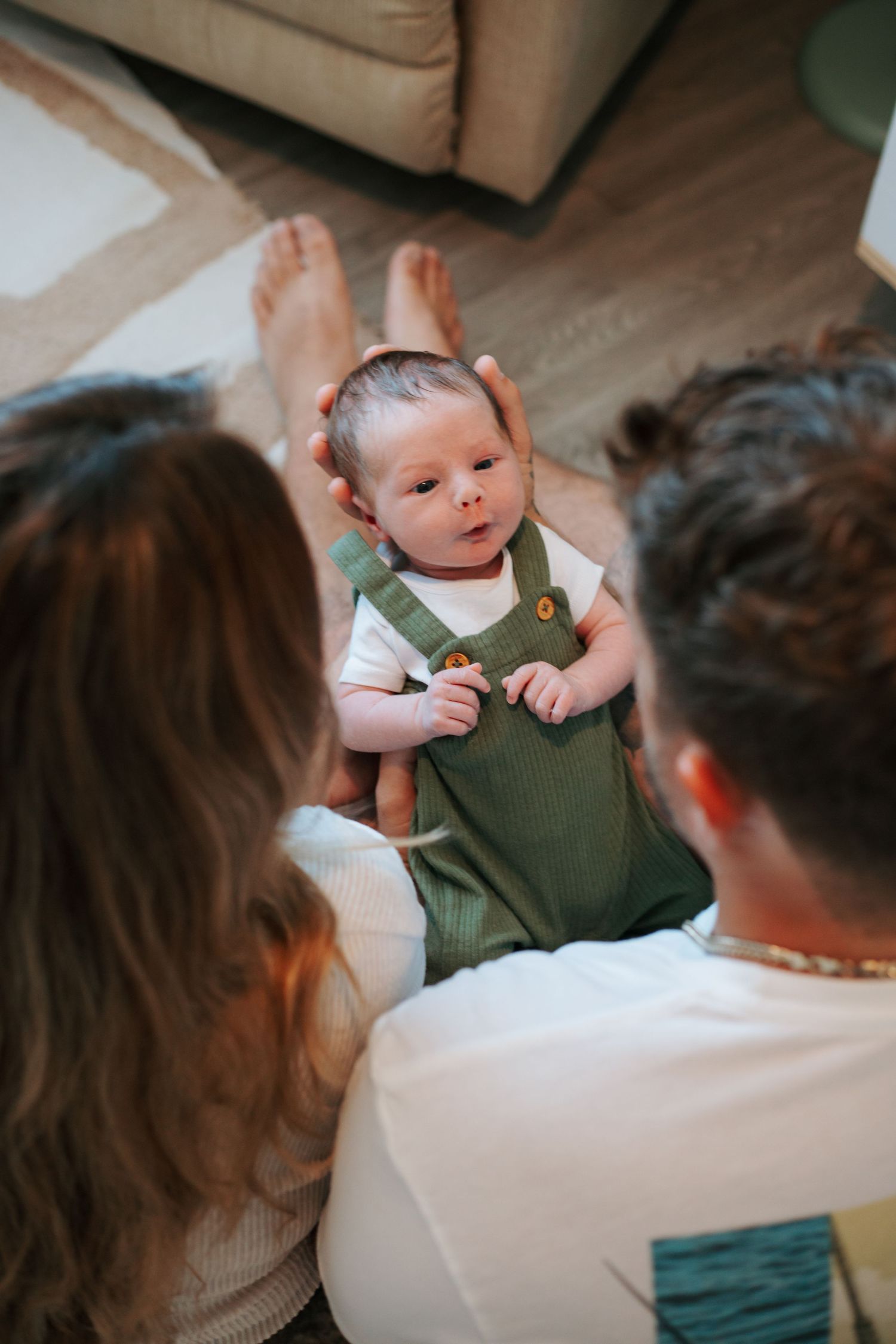 A baby wearing an olive green romper sits on a table while adults gather around in a casual indoor setting.