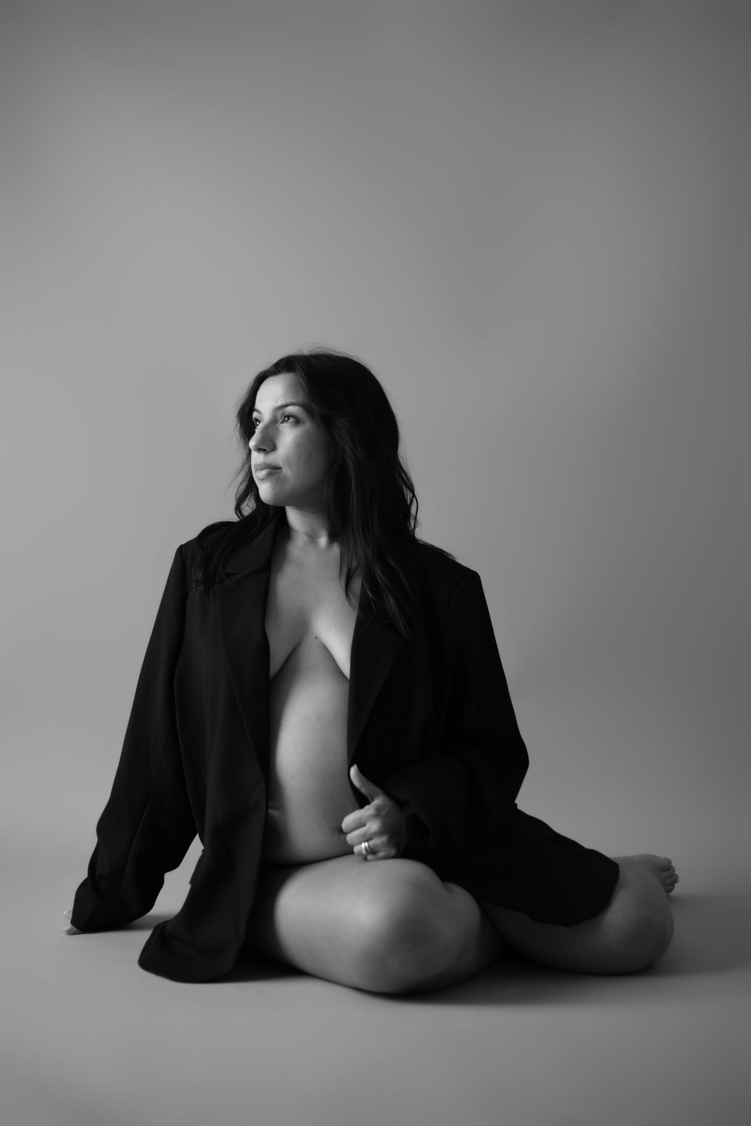 Artistic black and white boudoir photo of a person wearing an open black jacket in a minimalist studio setting.