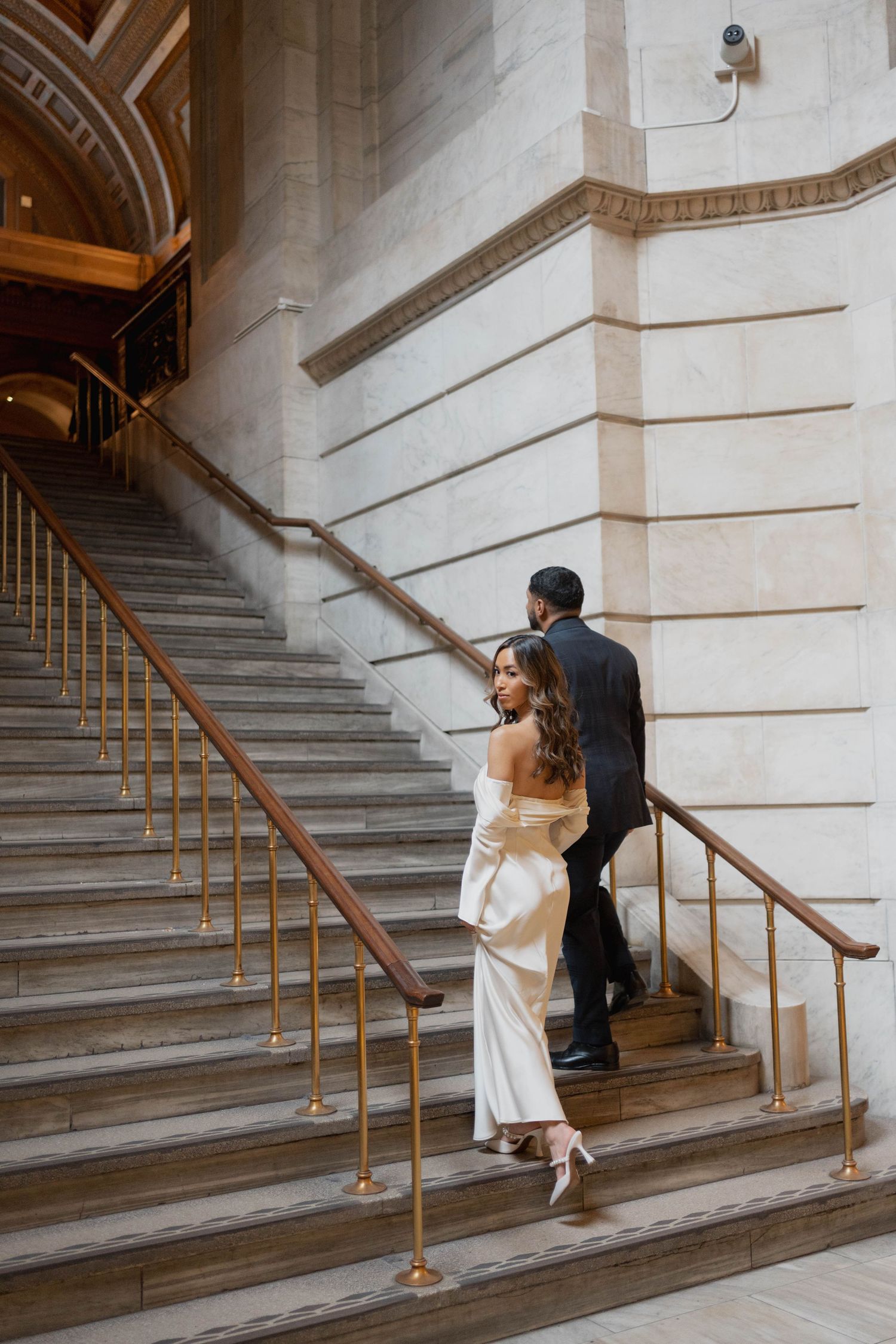 Couple walking up grand exterior staircase with gold railings, bride in white gown, groom in dark suit.
