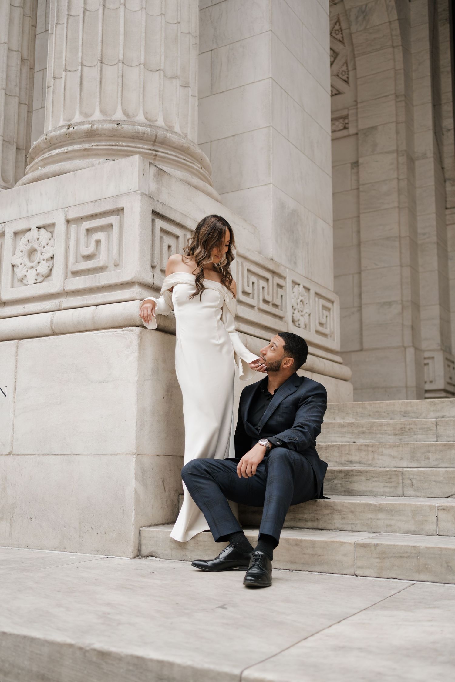 Groom sitting on steps gazing up at bride in white off-shoulder gown by stone column.