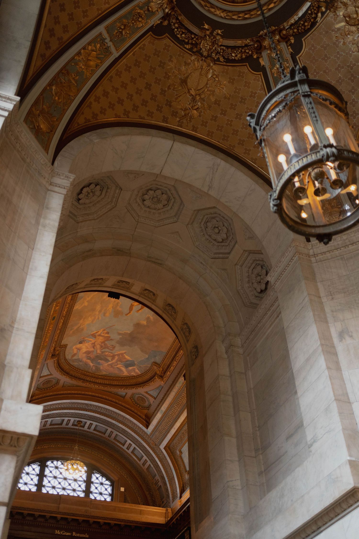 Ornate building interior featuring gilded ceiling, classical murals, arched doorways, and vintage chandelier.