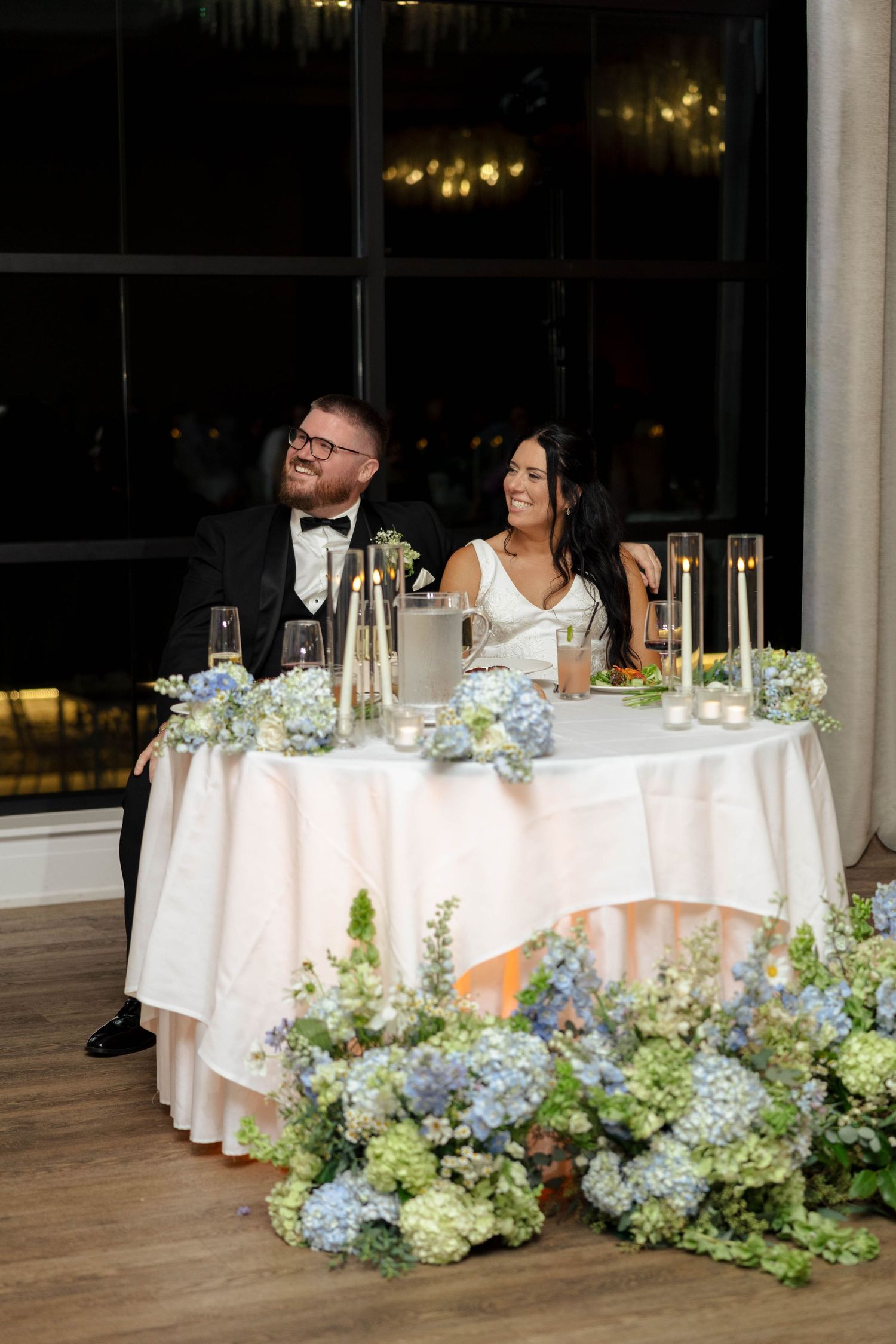 Bride and groom smiling at sweetheart table adorned with blue hydrangeas, candles, and white linens at wedding reception.