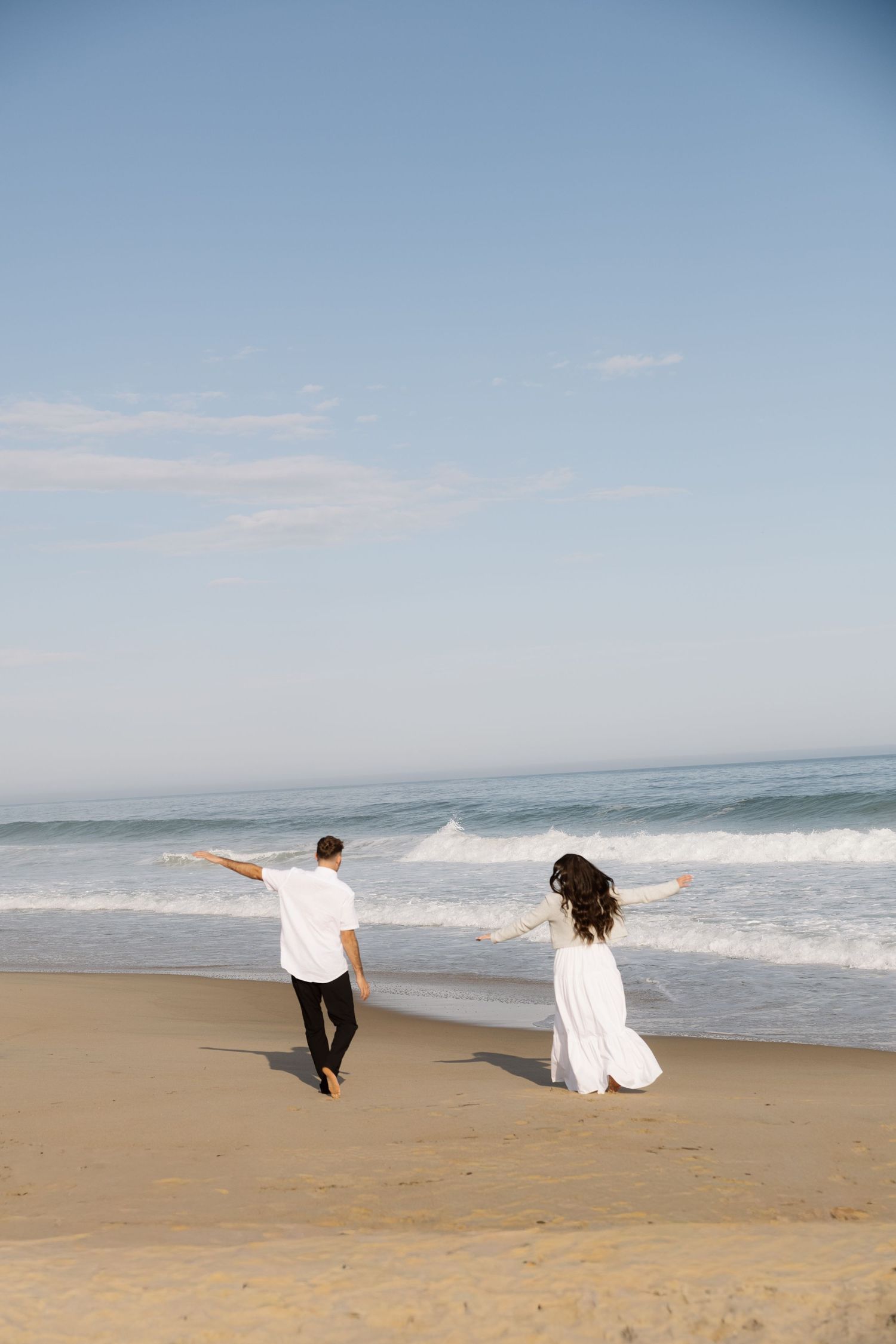 Couple in white wedding attire walking along a sandy beach with arms outstretched toward ocean waves.