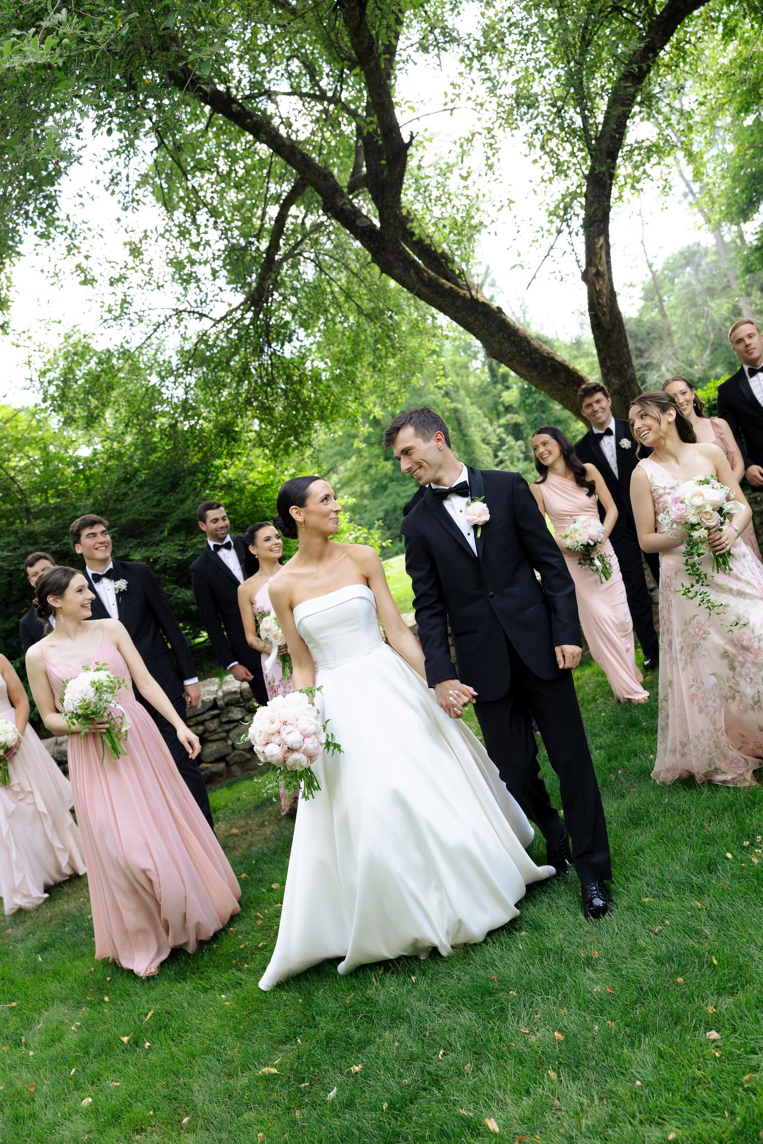 Bride and groom walk hand-in-hand outdoors, followed by wedding party in pink gowns and black tuxedos under lush green trees.