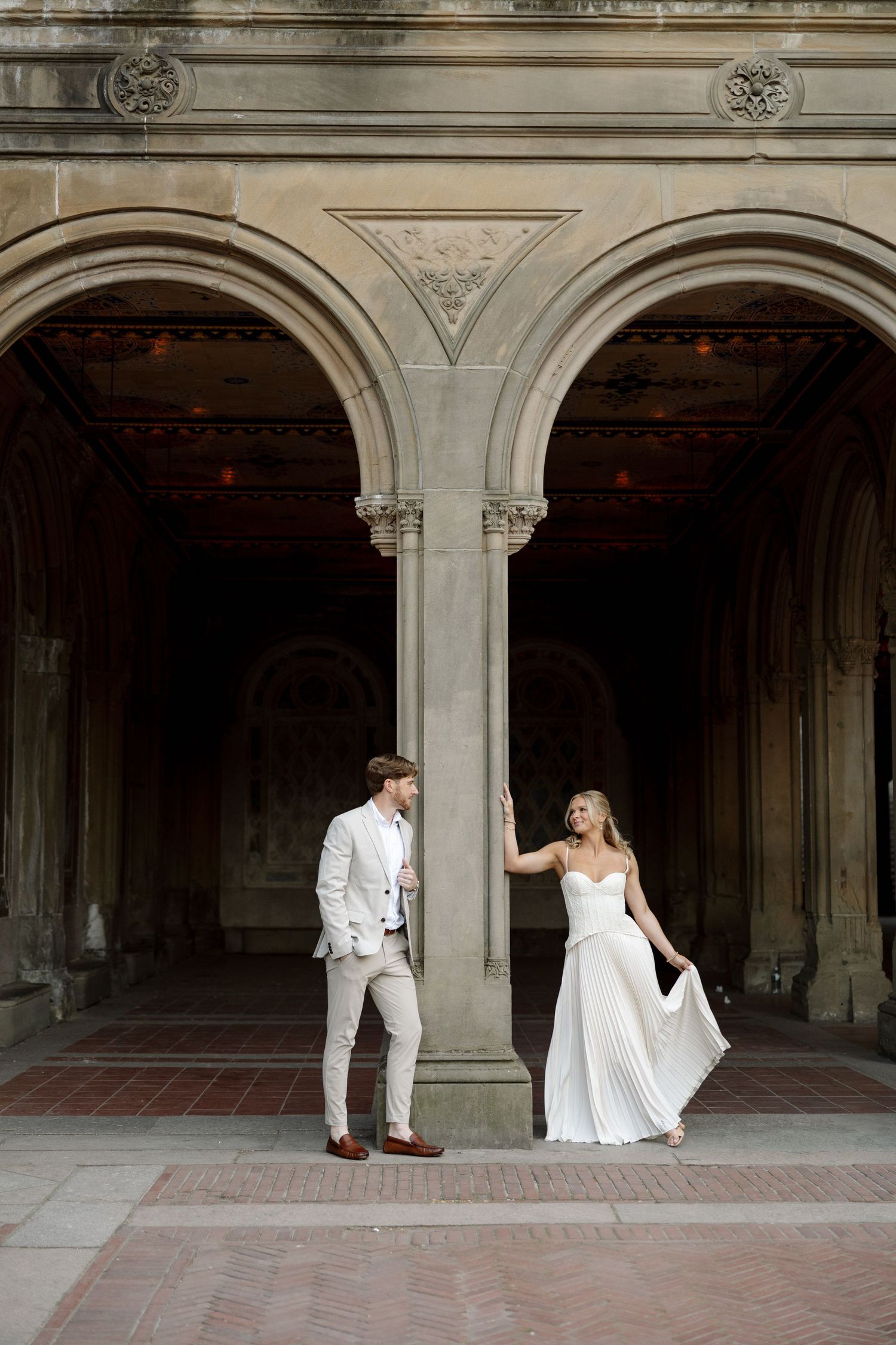 Central Park New York Photography - Bride in flowing white pleated gown and groom in beige suit holding hands under stone arched colonnade.