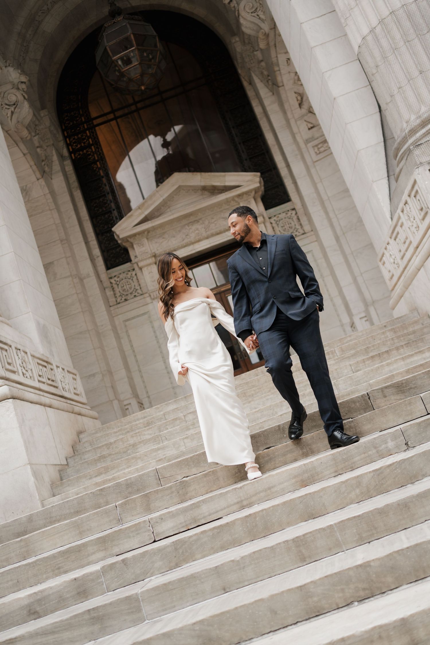 Bride and groom holding hands walking down grand marble steps of neoclassical building.