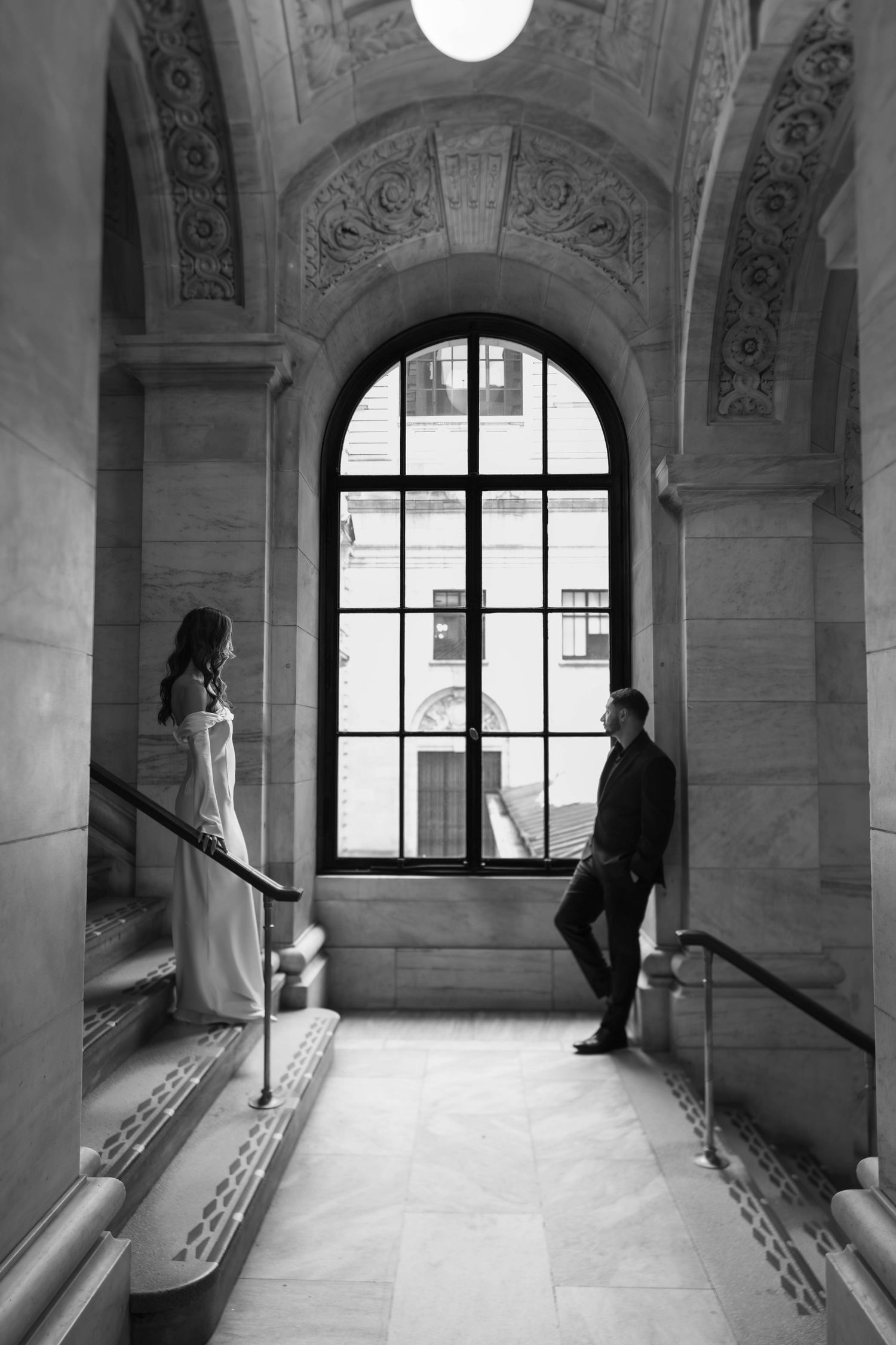 Black and white photo of bride and groom standing apart on marble stairs beneath grand arched window.