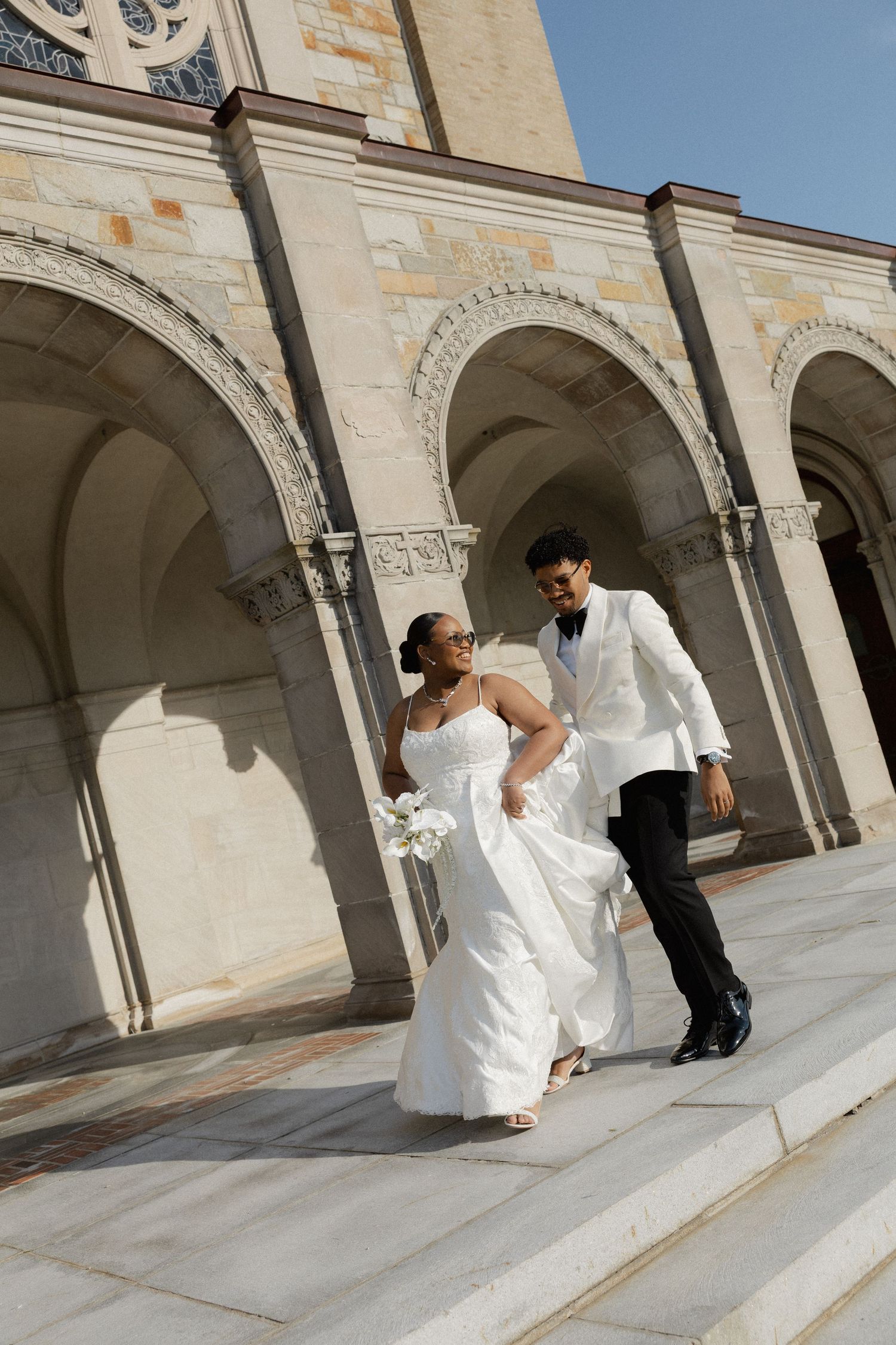 Bride and groom walking down steps outside elegant stone church with arched columns on their wedding day.