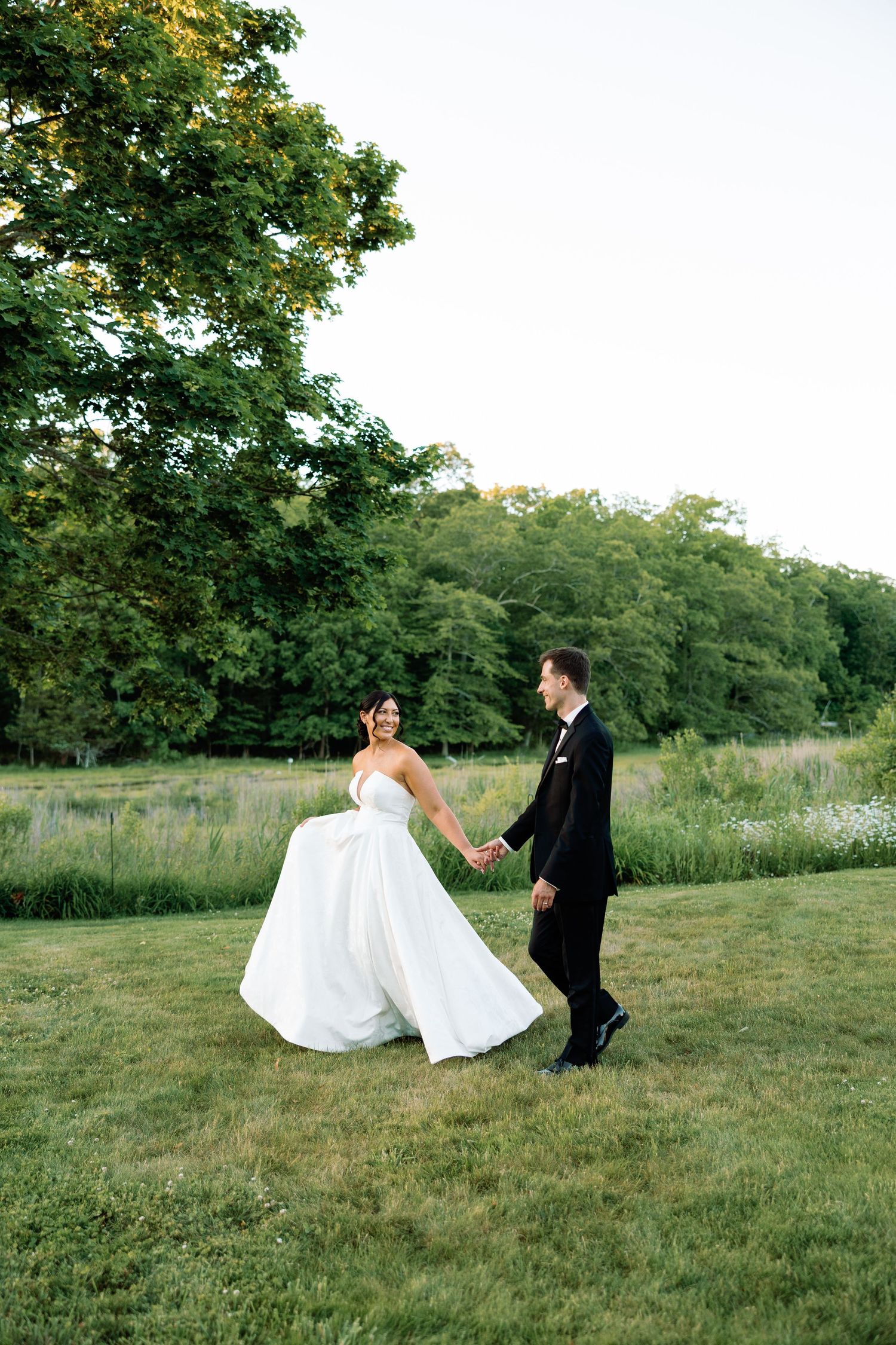 Lord Thompson Manor - CT Wedding Photography - bride lifts gown while holding grooms hand walking across lawn at sunset.