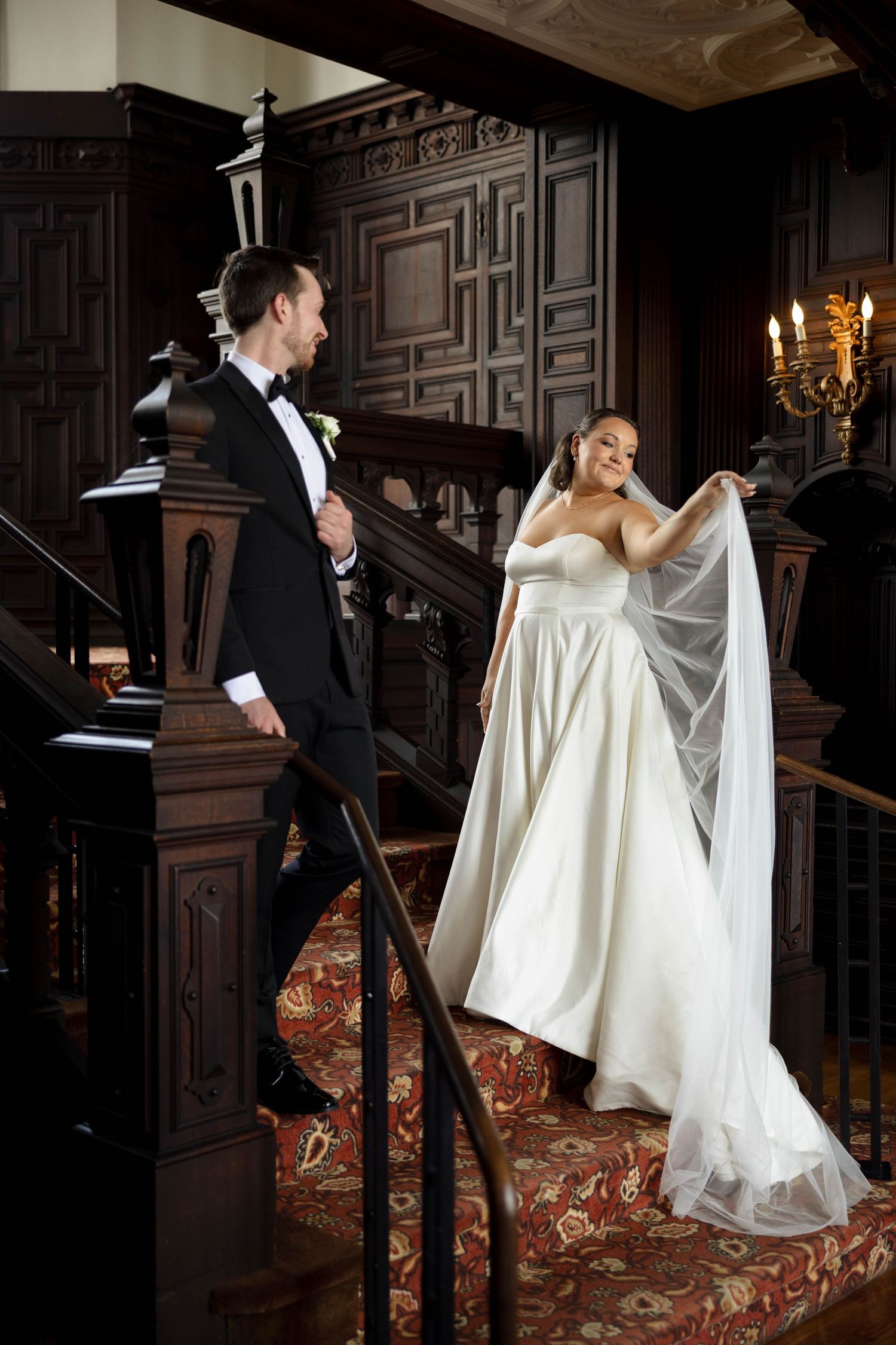 Bride in white strapless gown holds veil on ornate dark wood staircase beside groom in black tuxedo.