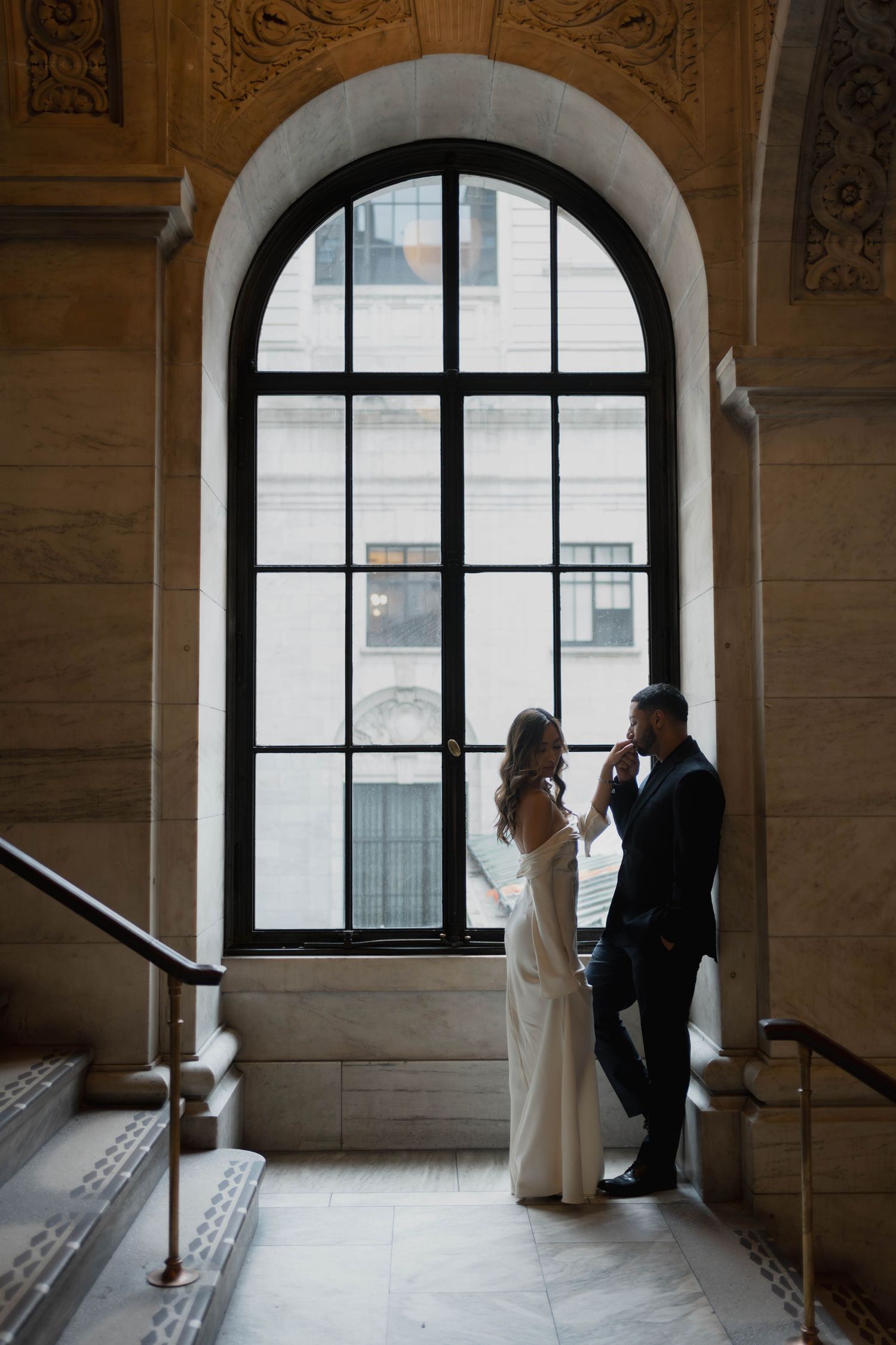 Bride and groom sharing tender moment beneath large arched window in elegant marble building interior.