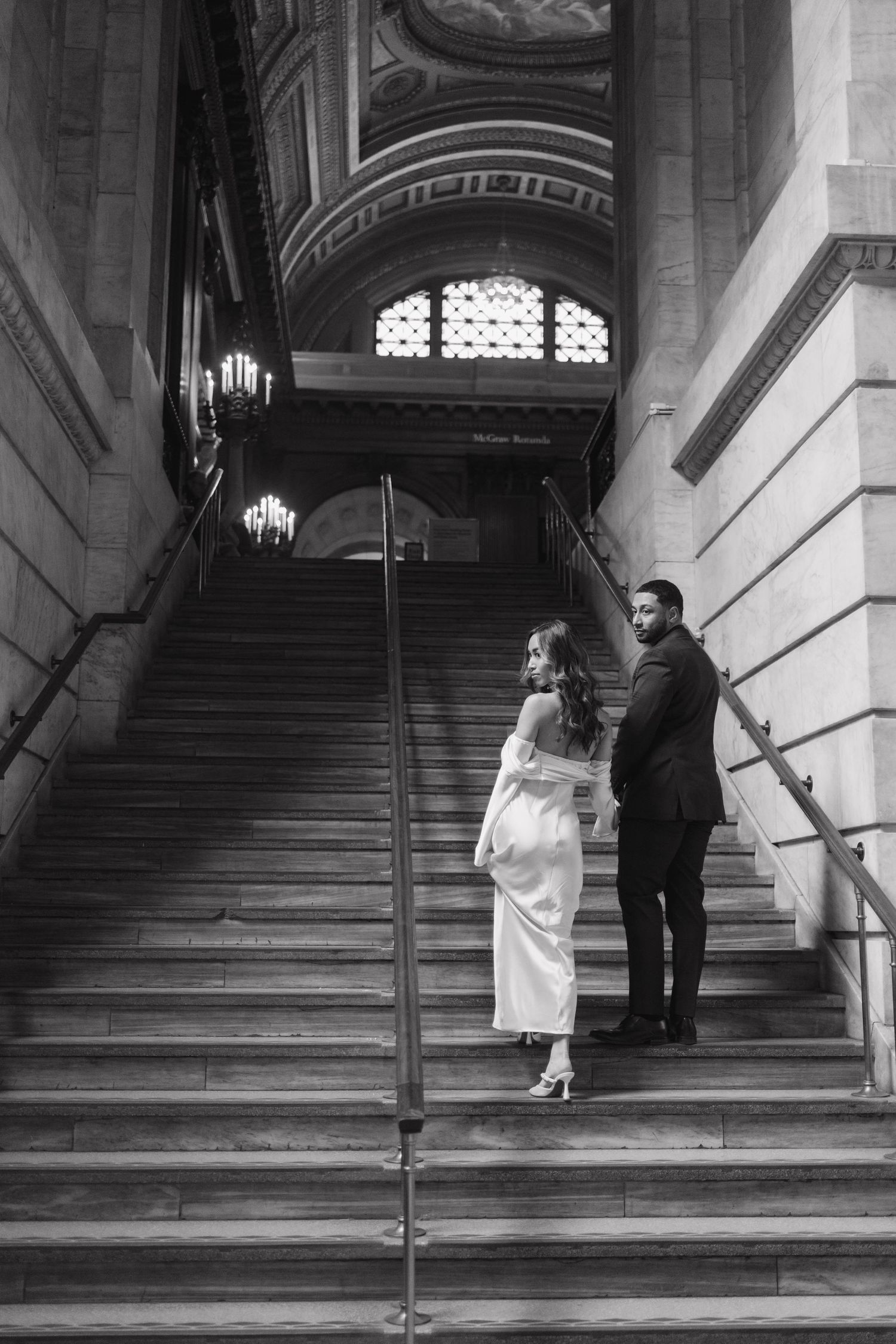 Black and white photo of couple on grand staircase beneath ornate gilded ceiling and chandelier.