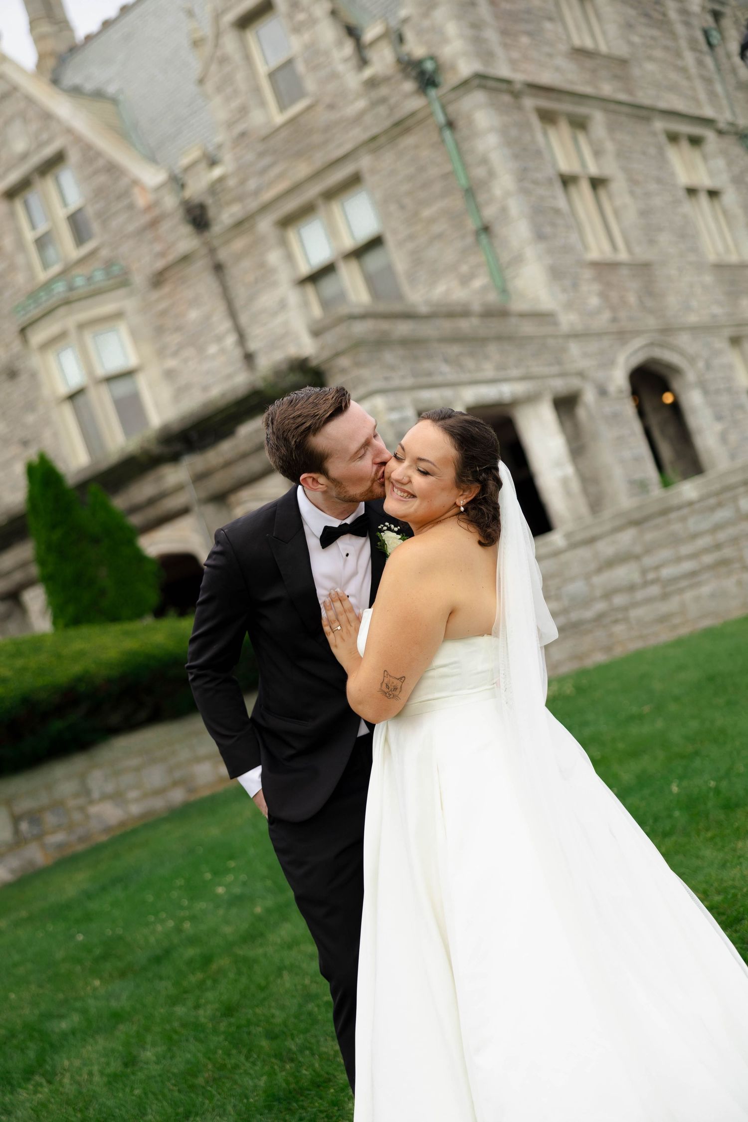 Groom in black tuxedo kisses bride in white strapless gown outside a stone castle venue on their wedding day.
