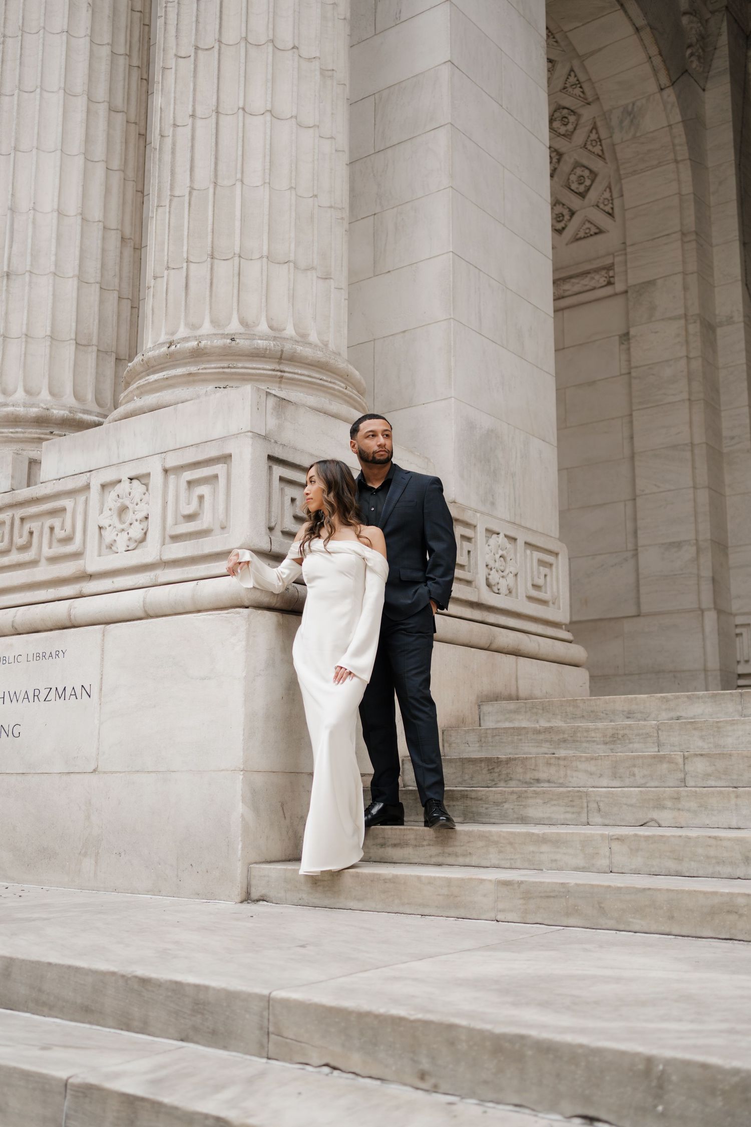 Bride and groom standing together on steps beside engraved stone column of library.