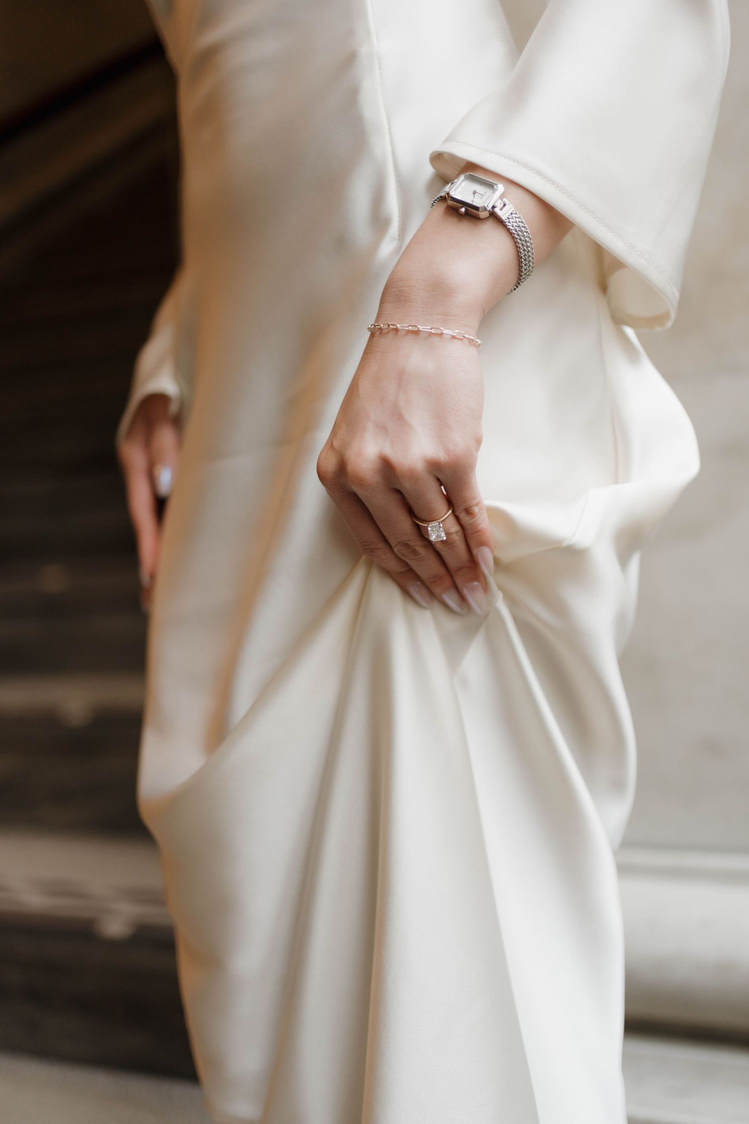 Close-up of bride's hand showing diamond engagement ring, bracelet, and watch against white satin gown.