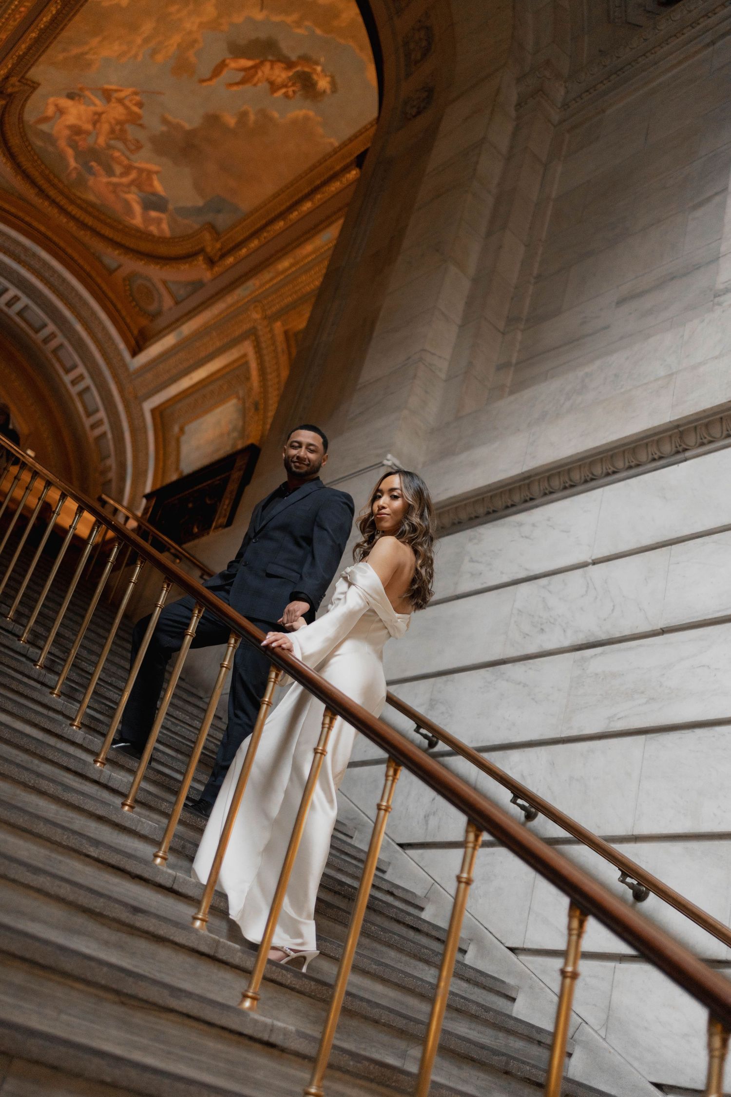 Bride in white gown and groom in dark suit pose on elegant staircase beneath golden painted ceiling.