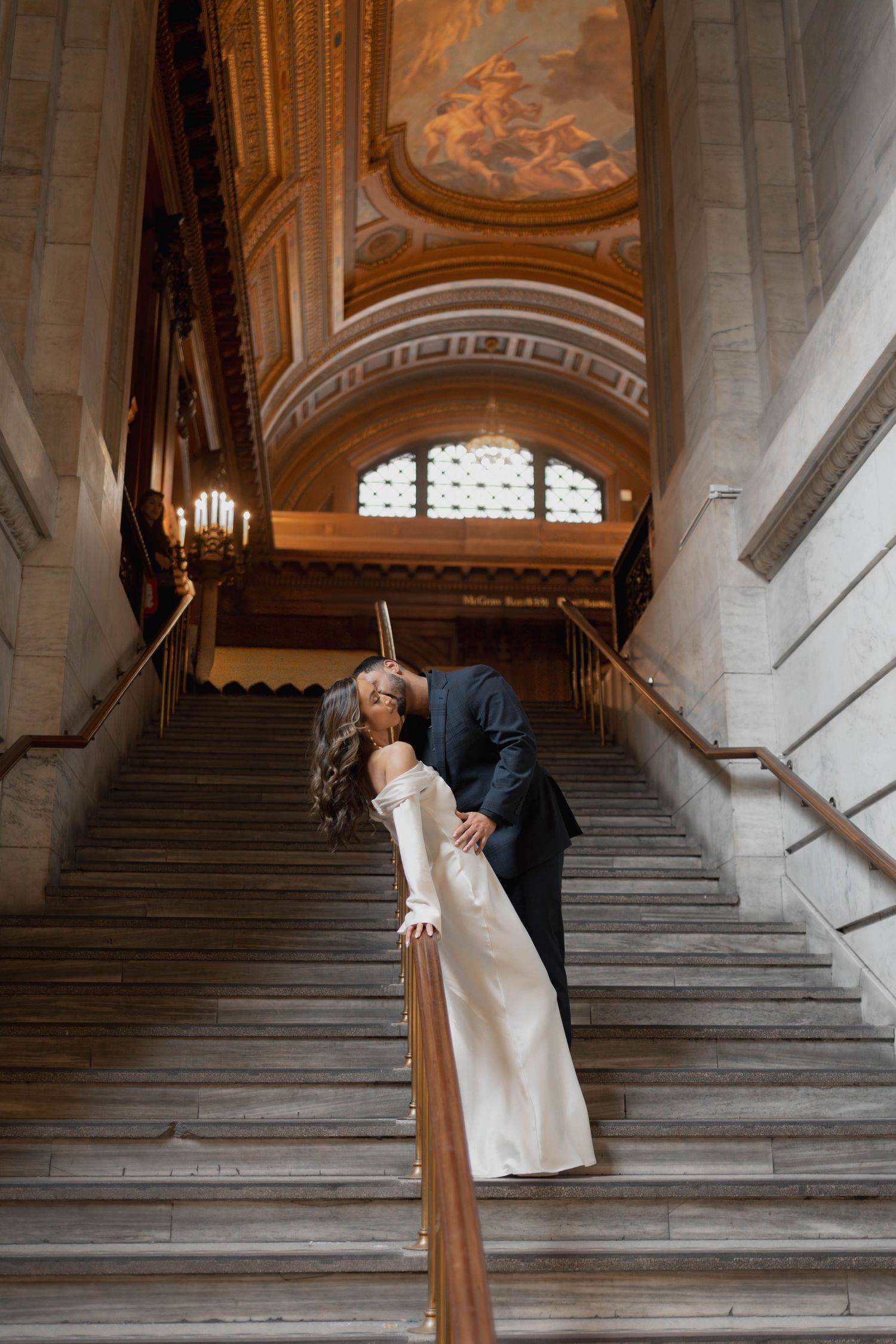 Bride and groom share a romantic kiss on grand marble staircase with ornate painted ceiling above.