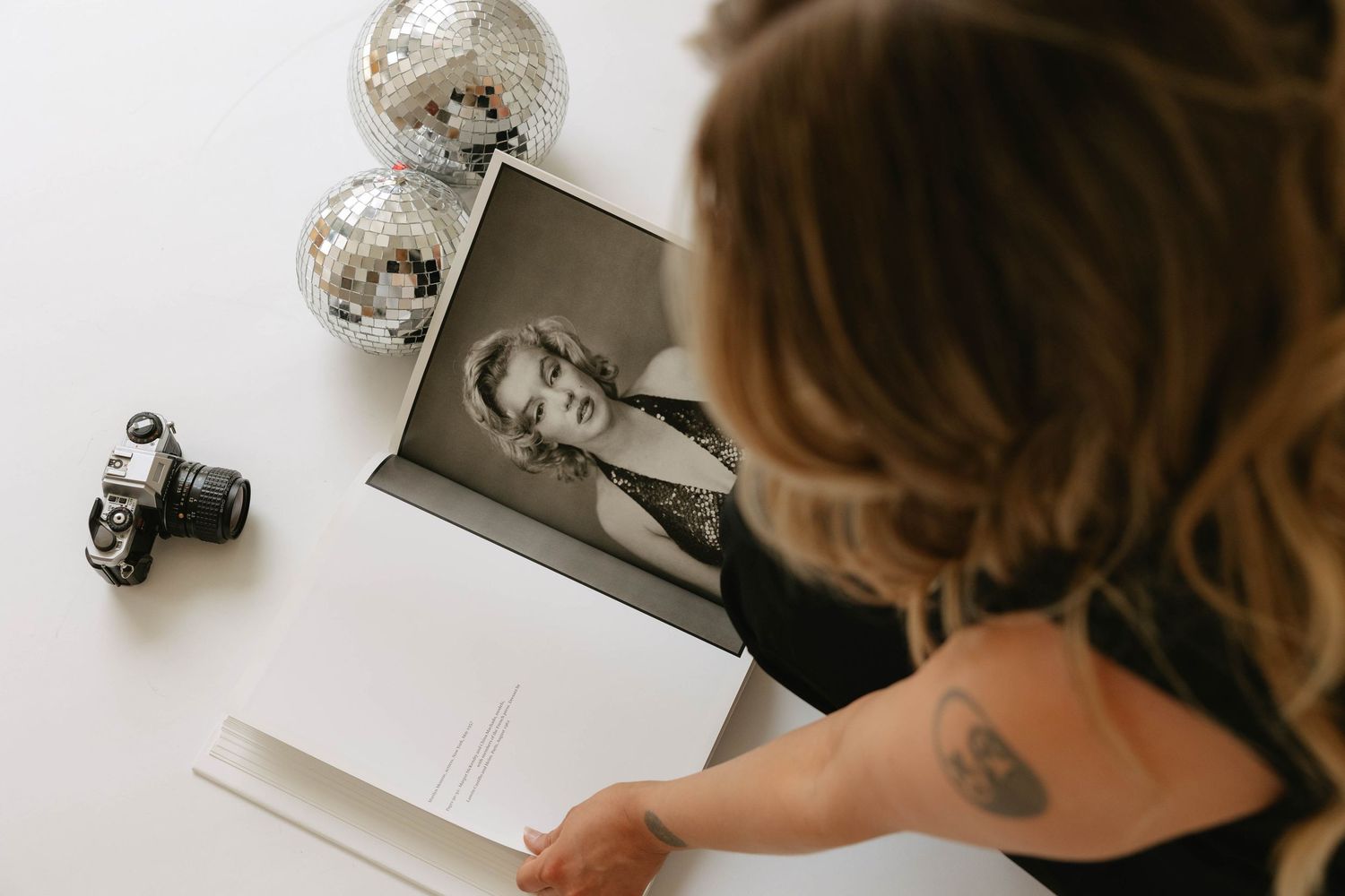 Woman with tattoo reviewing black and white portrait photo on white desk with disco balls and film camera.