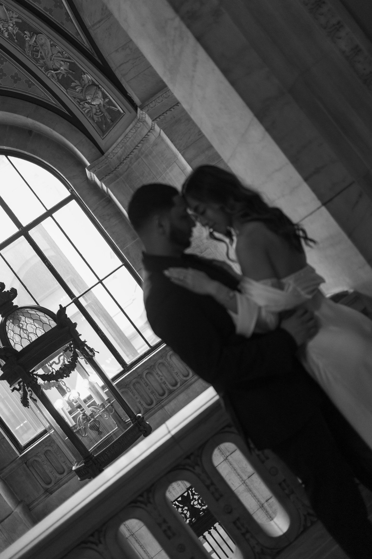 Black and white photo of bride and groom embracing near arched window in elegant marble building.