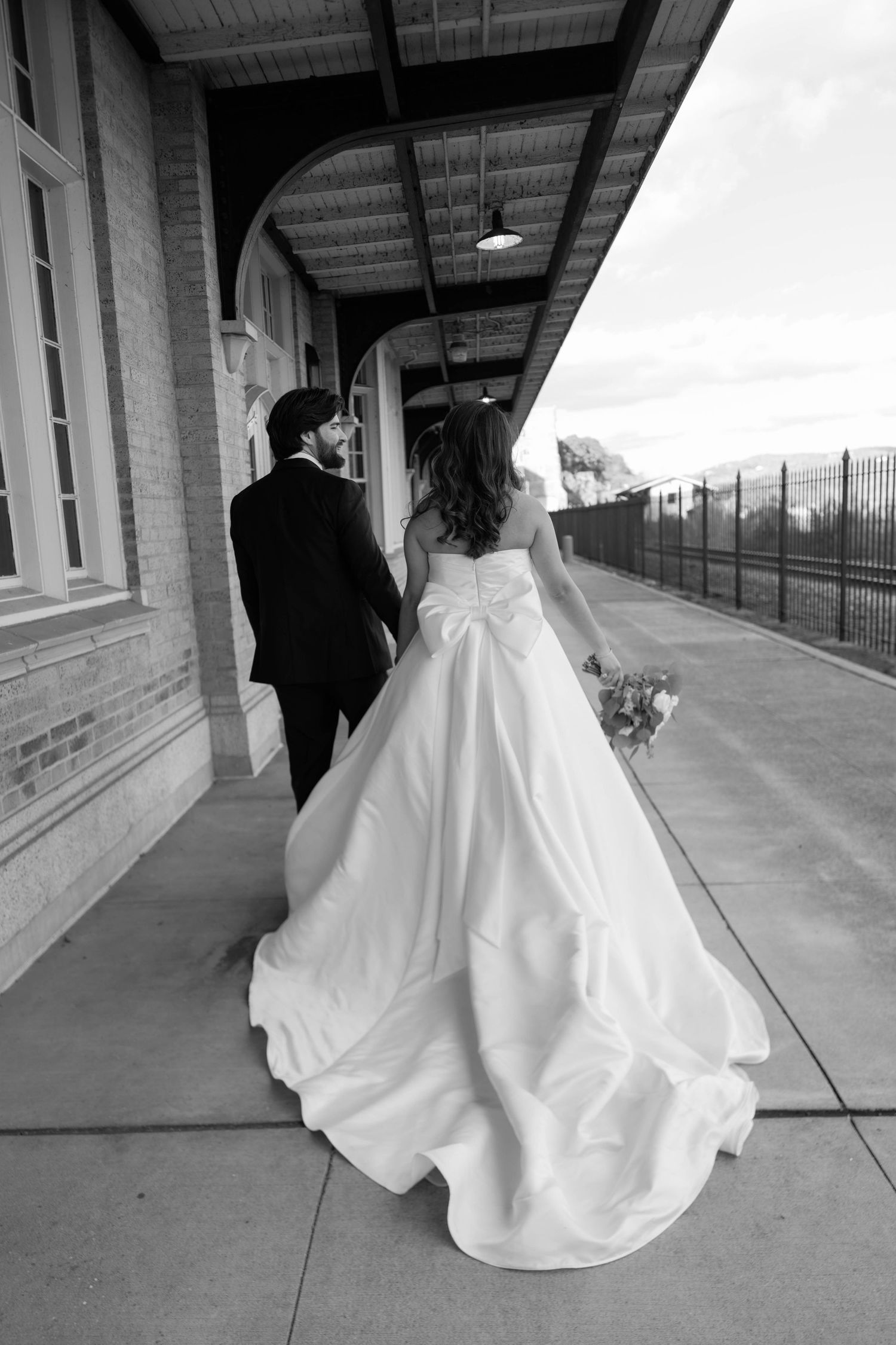 Black and white photo of bride and groom walking hand in hand along a covered brick train station platform.