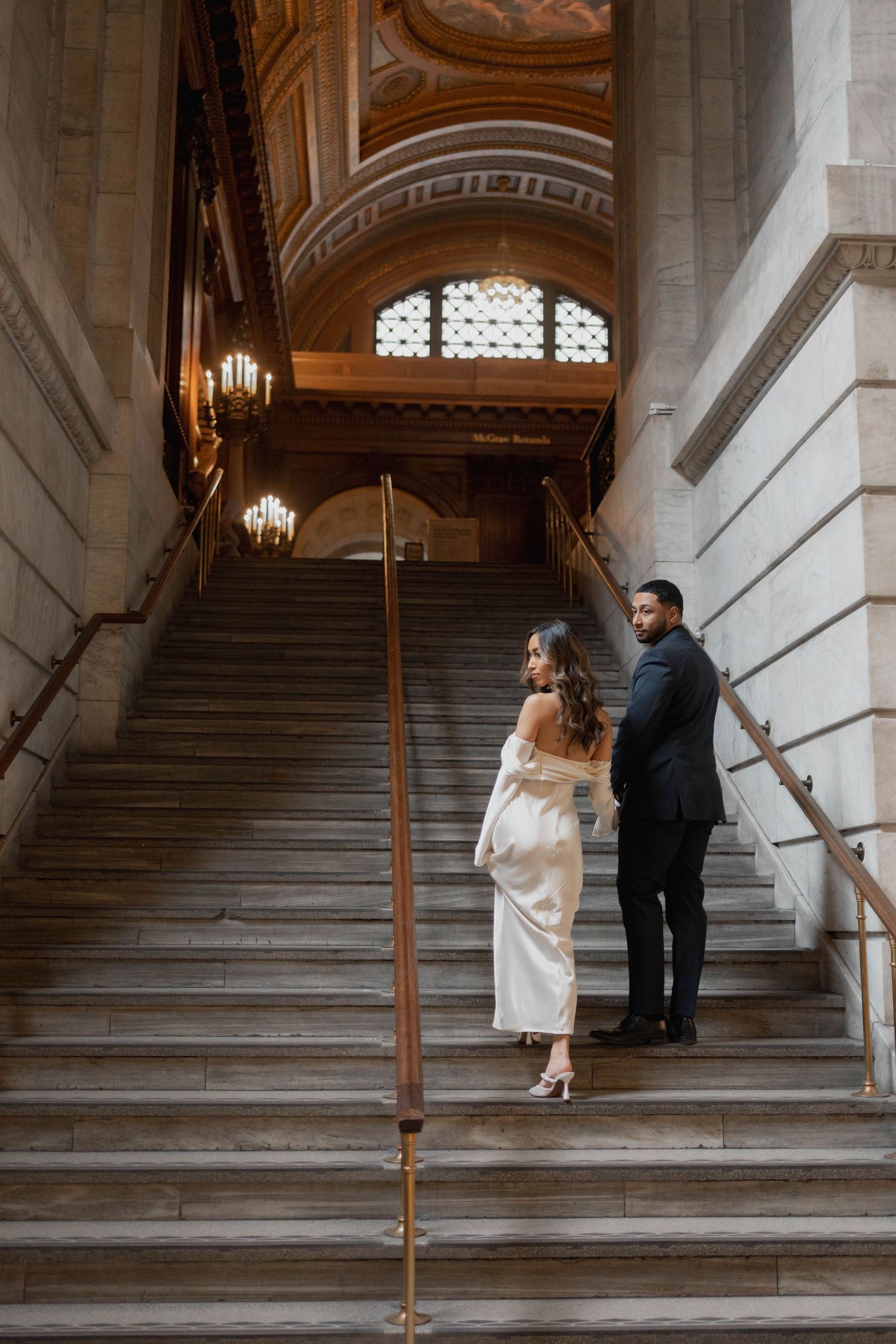 Couple in wedding attire walking up grand marble staircase in ornate historic building with chandeliers.