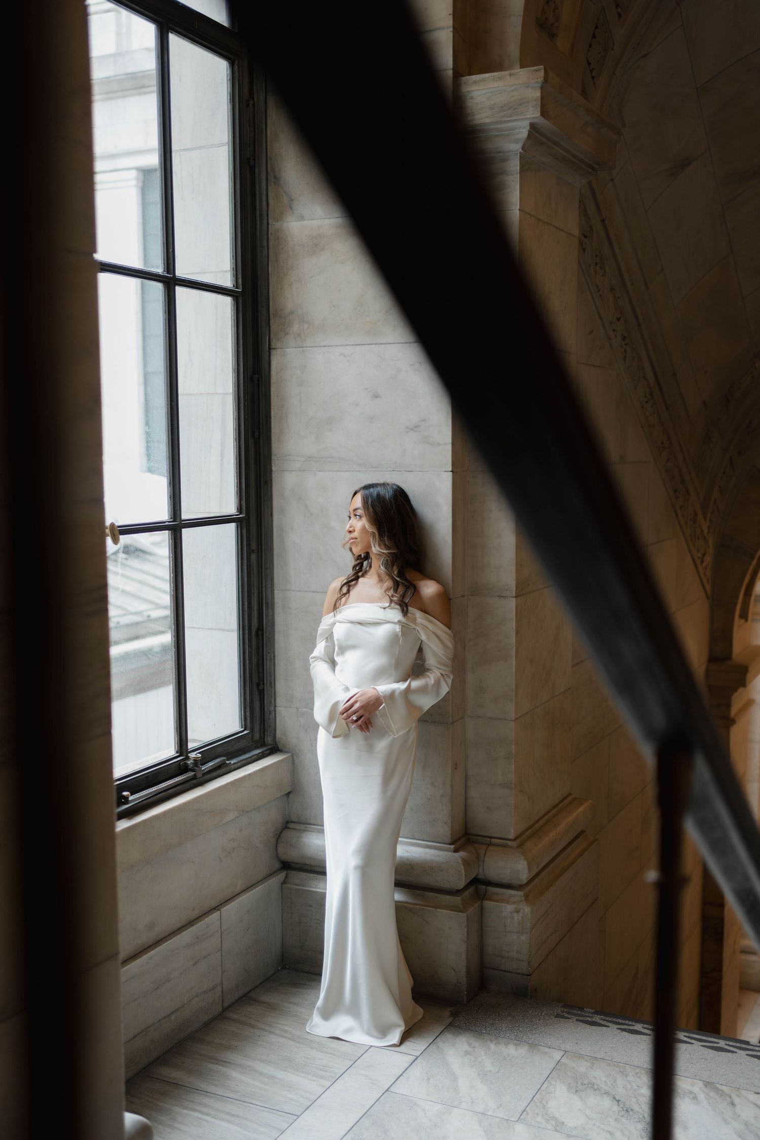 Bride in white off-shoulder long sleeve gown standing by tall window in ornate marble building.