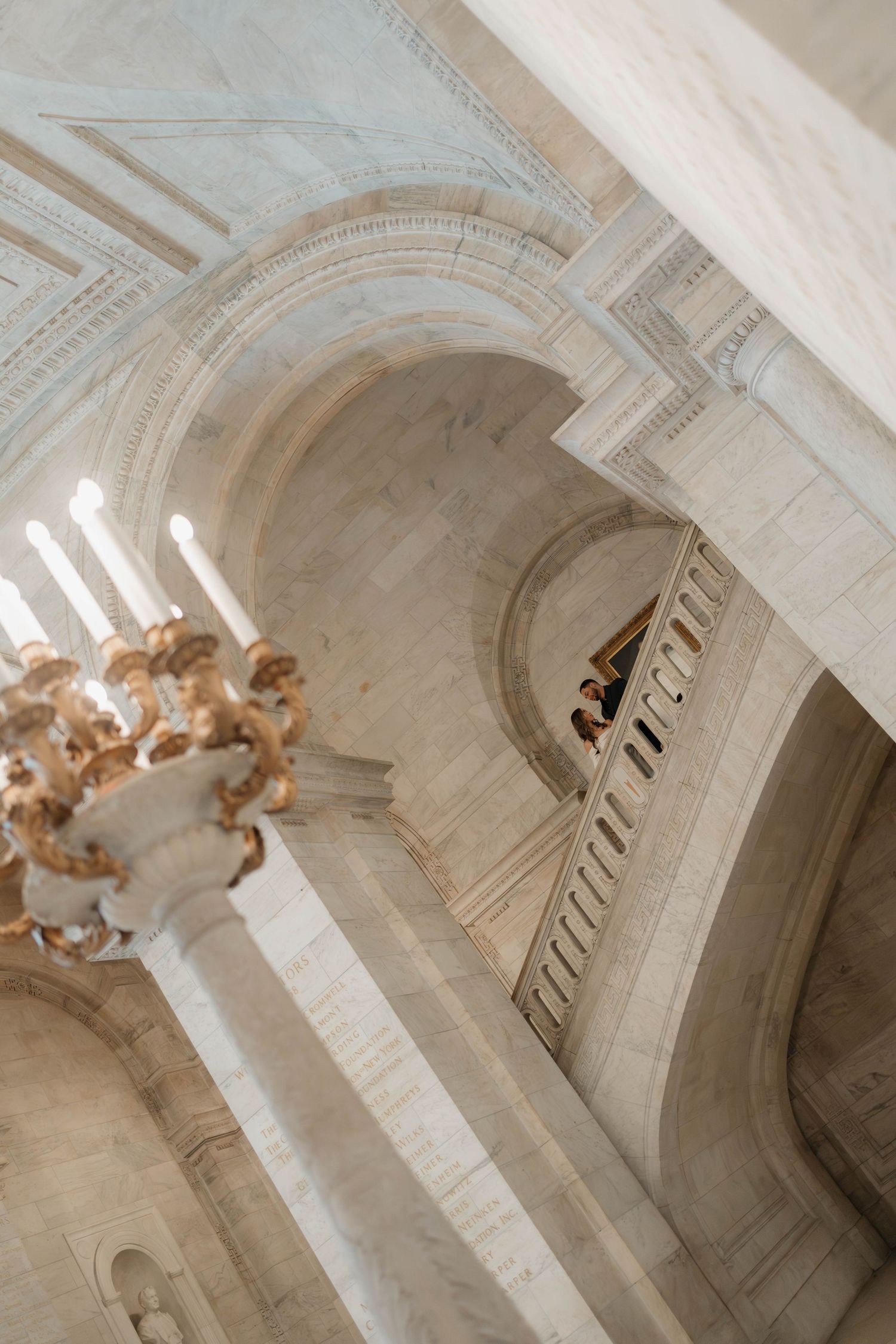 Couple standing on an upper balcony of the New York Public Library amid soaring marble arches.