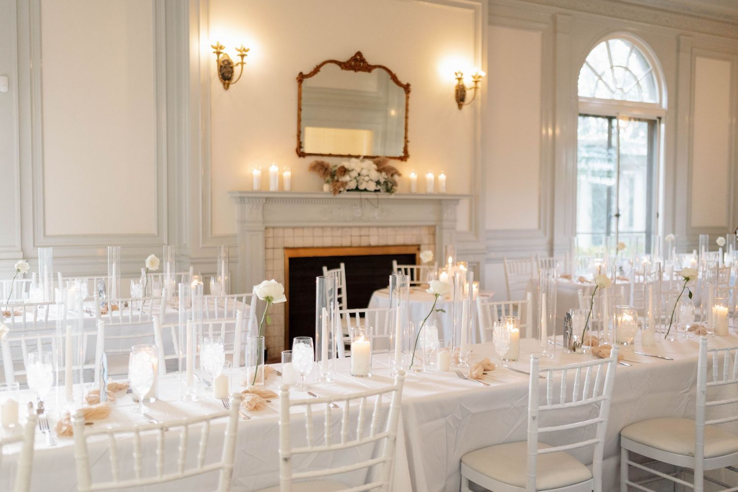Elegant white wedding reception hall with chiavari chairs, candlelit tables, fireplace, and ornate mirror in a classic venue.