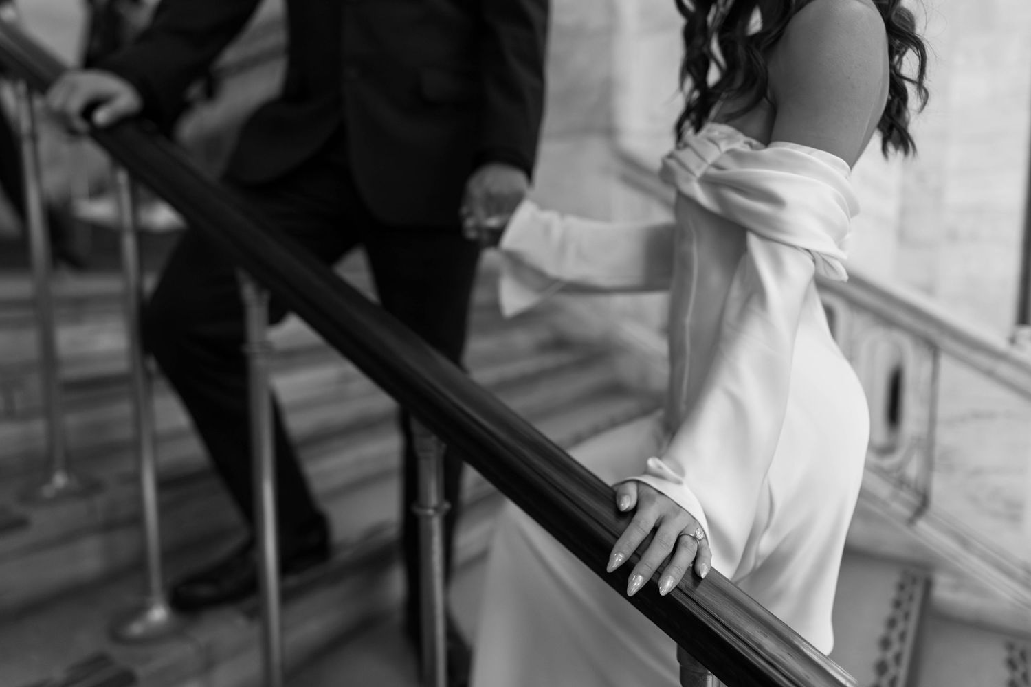 Black and white close-up of a bride in an off-shoulder gown holding a staircase railing.