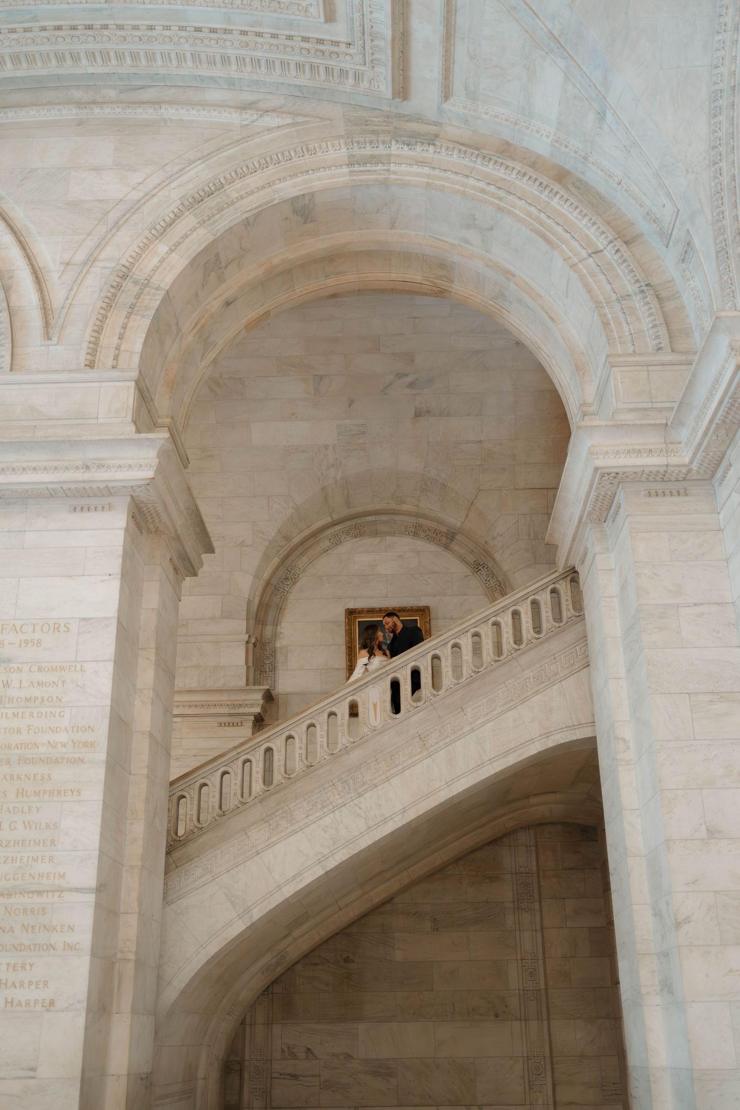 Wedding couple posed on a sweeping marble staircase beneath grand arched ceilings of a historic library.
