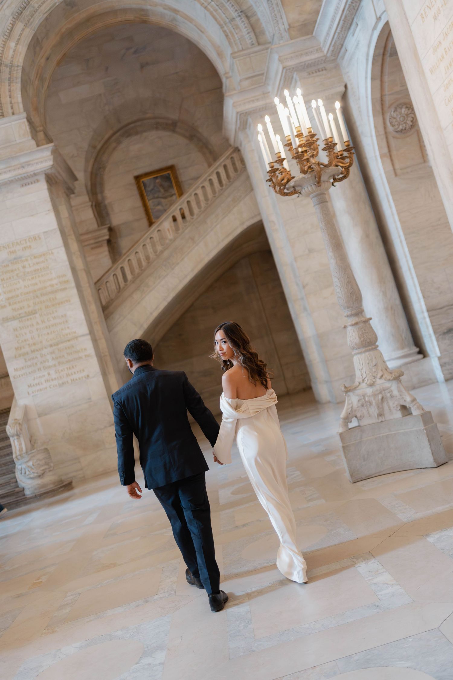Bride in white gown and groom in dark suit walking through the grand marble halls of a historic library.