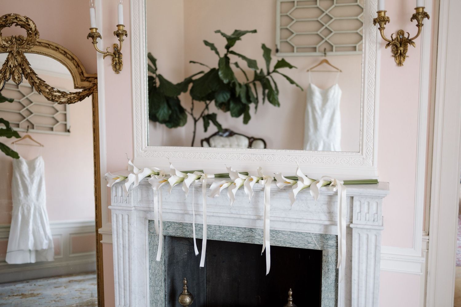 Elegant bridal suite with white wedding dress hanging over a marble fireplace in a pink room with gold mirrors.