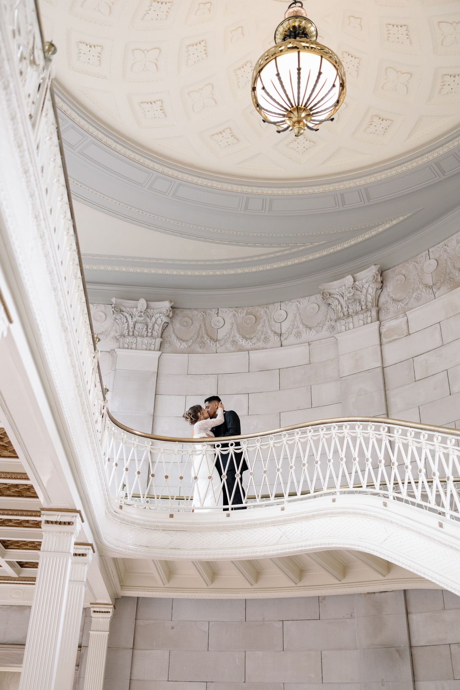 Bride and groom sharing romantic moment on curved white balcony beneath ornate domed ceiling with chandelier.