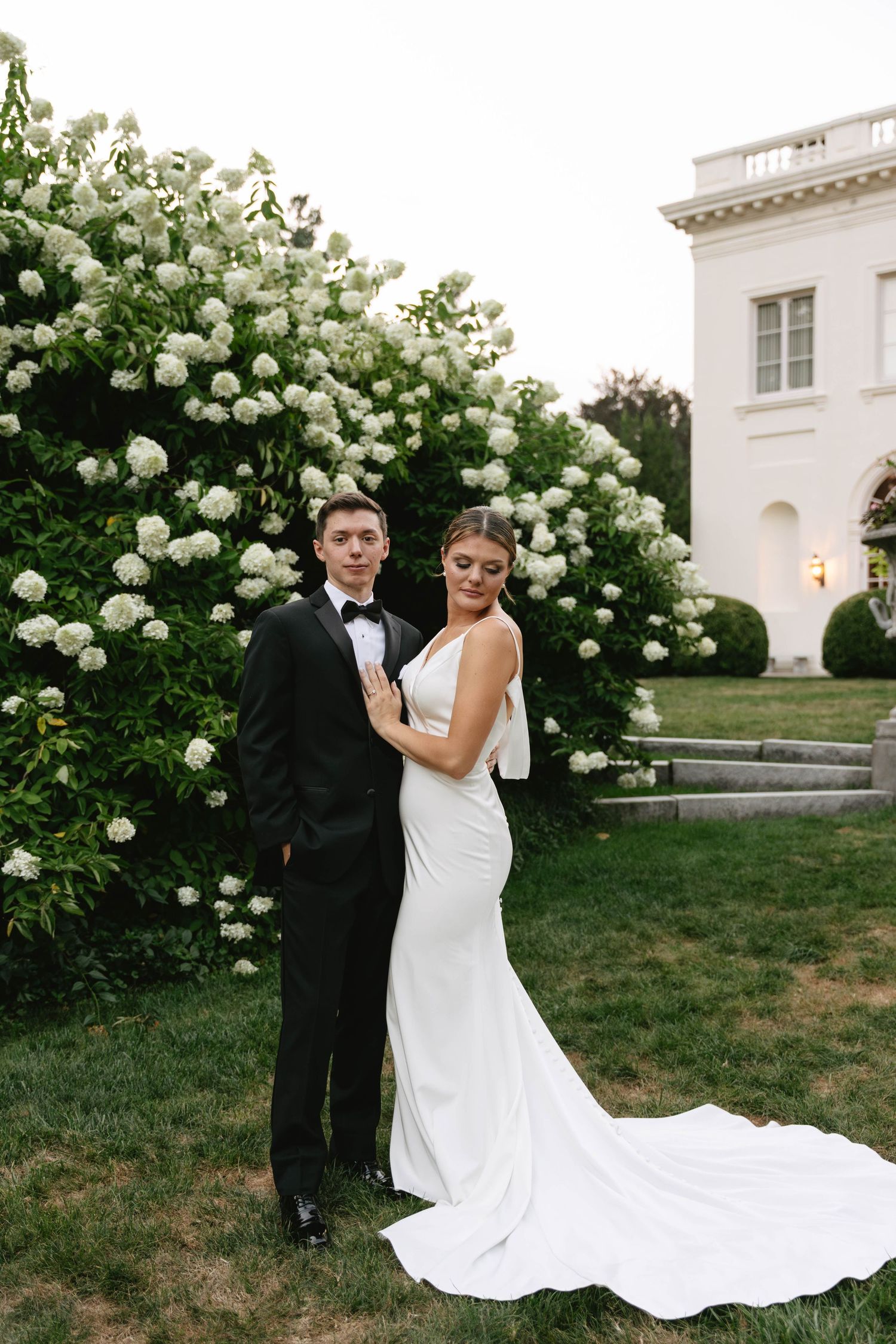 Bride in sleek white gown and groom in tuxedo posing before blooming white hydrangea bushes.