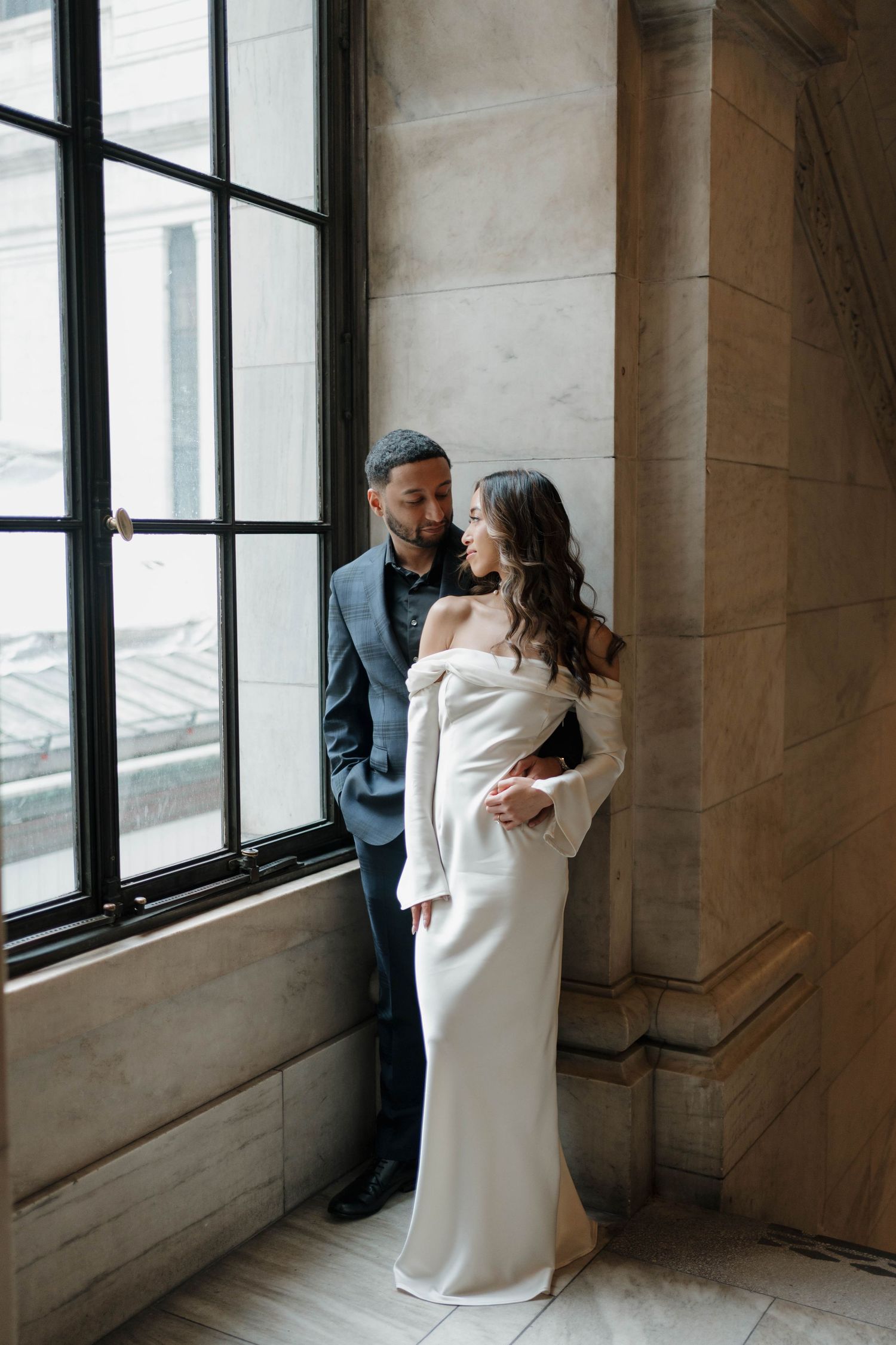 Couple sharing intimate moment by tall window, bride in white off-shoulder gown, groom in dark suit in ornate building.