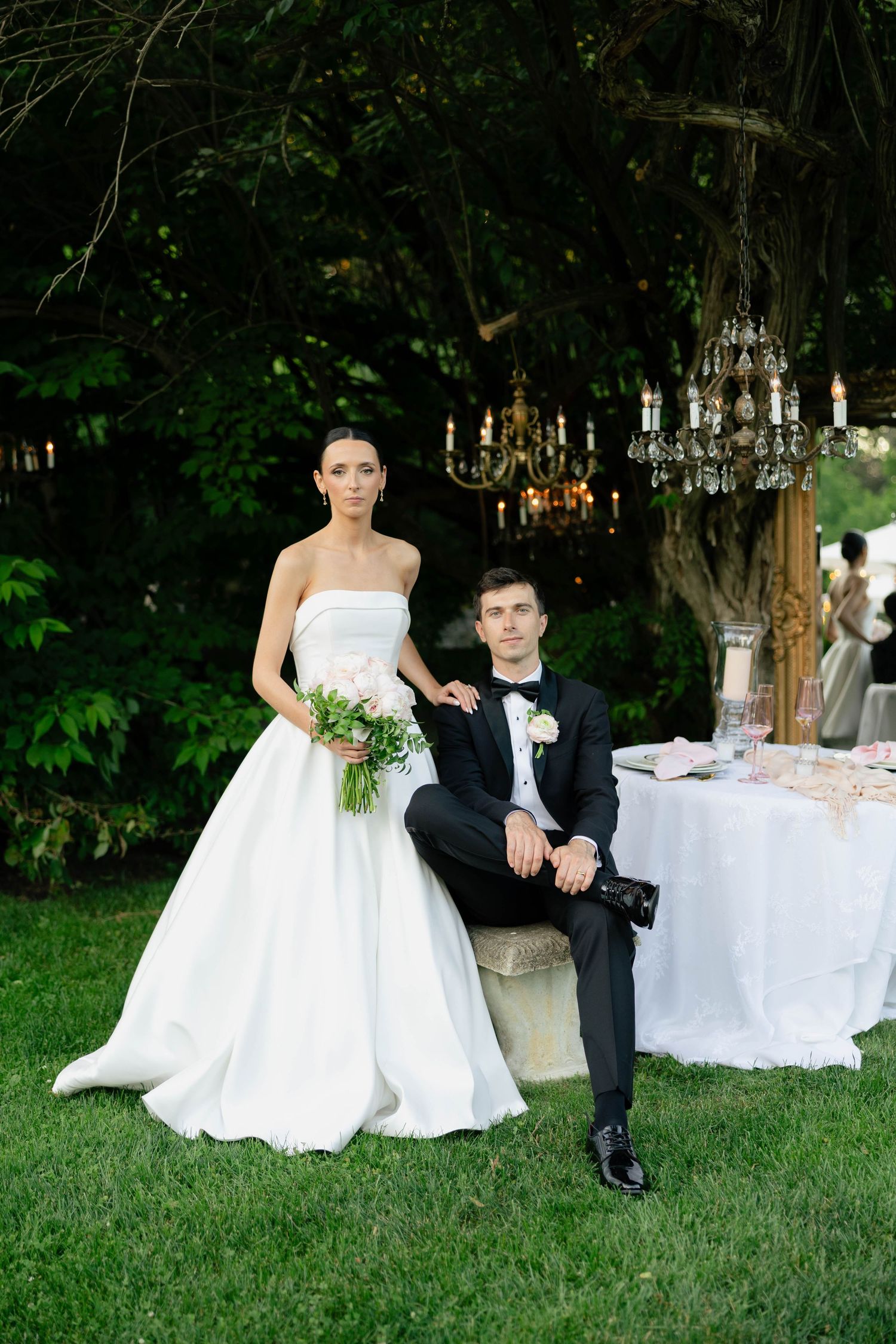 Elegant bride in strapless white gown holding bouquet stands beside groom in black tuxedo seated outdoors near chandelier.
