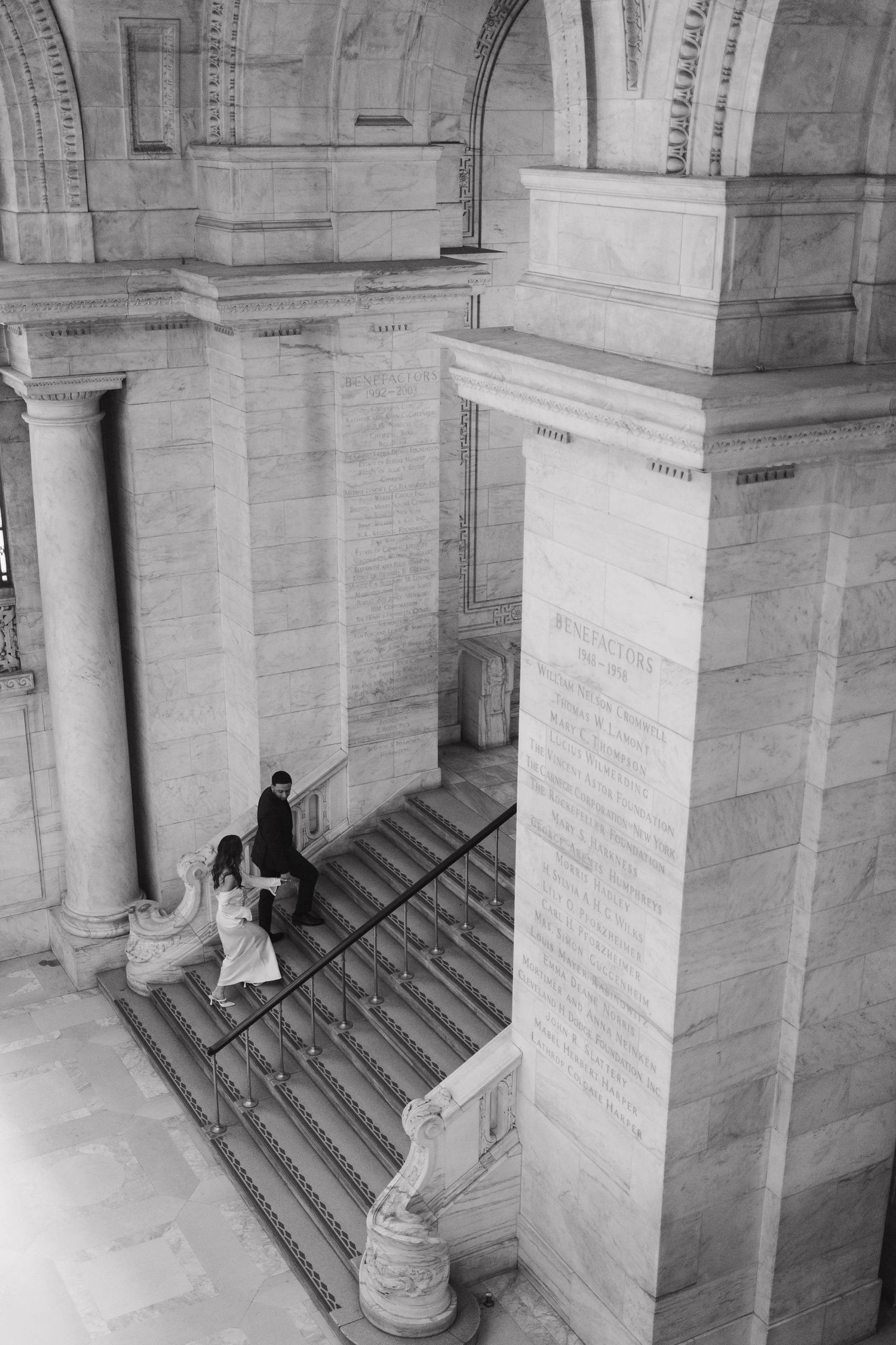 Black and white aerial view of a wedding couple ascending grand marble stairs in a historic building.